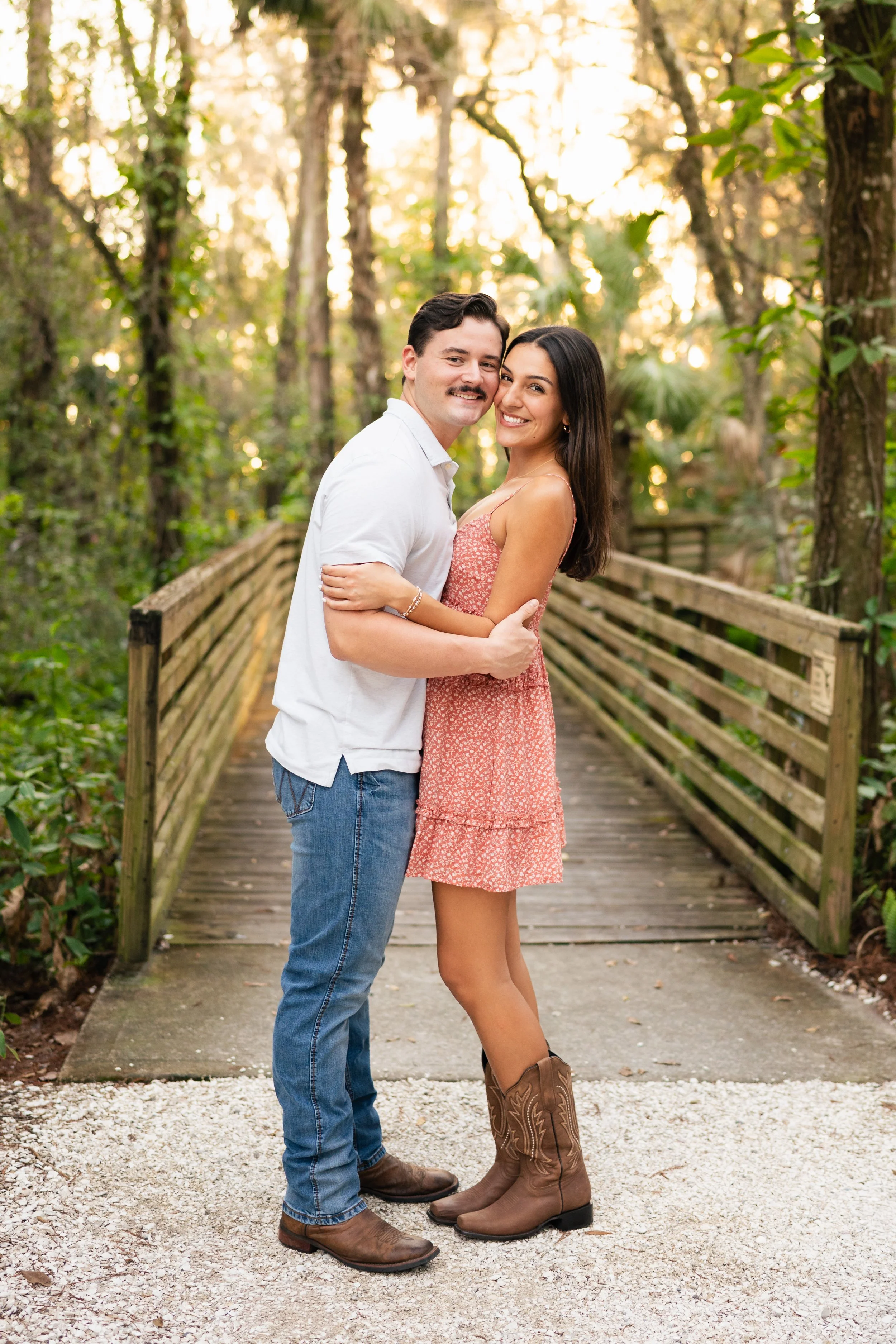 A smiling couple embracing on a wooden bridge in a wooded park during sunset.