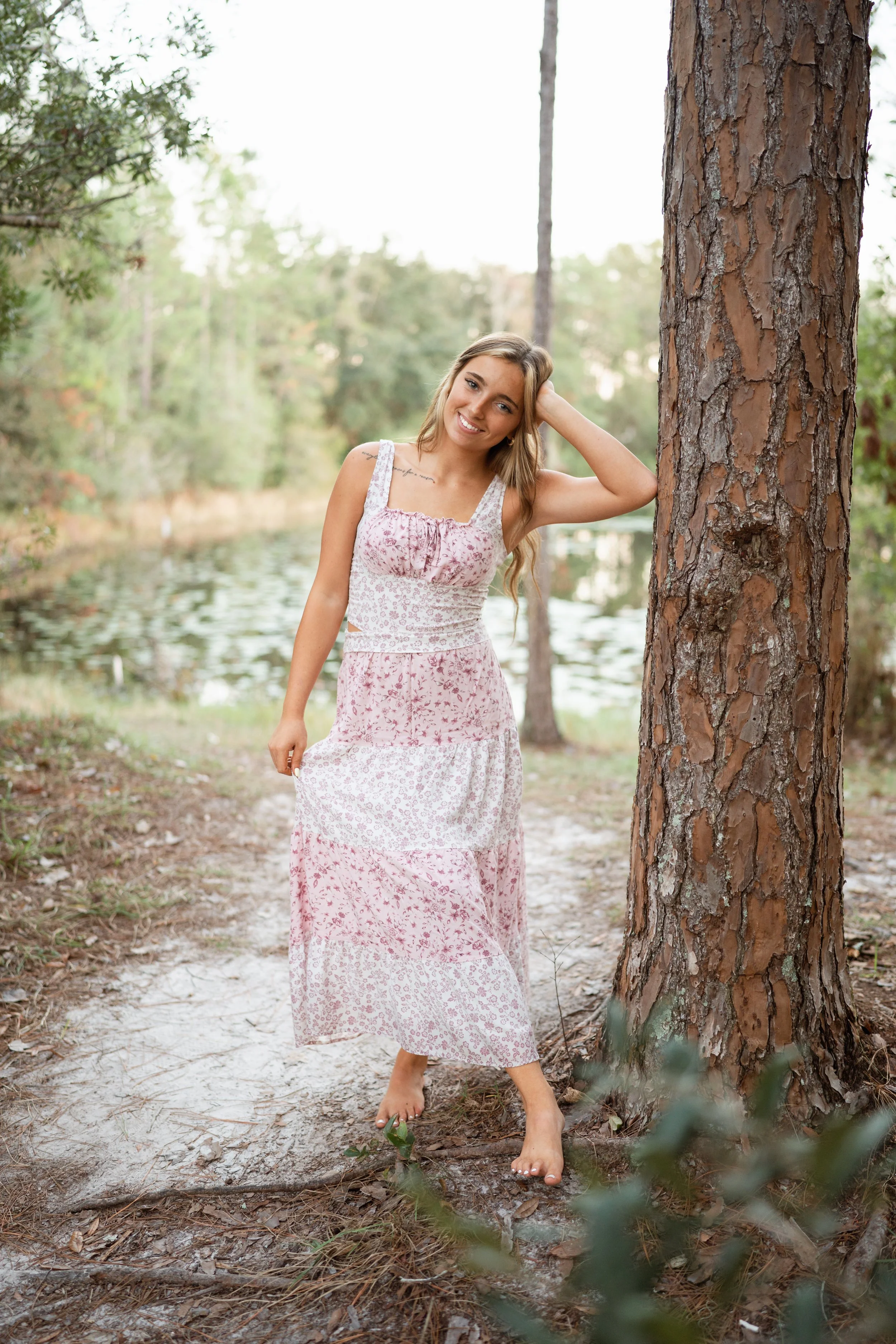 A young woman with long blonde hair standing outdoors on a forest path near a lake, smiling, barefoot, wearing a pink and white floral dress.