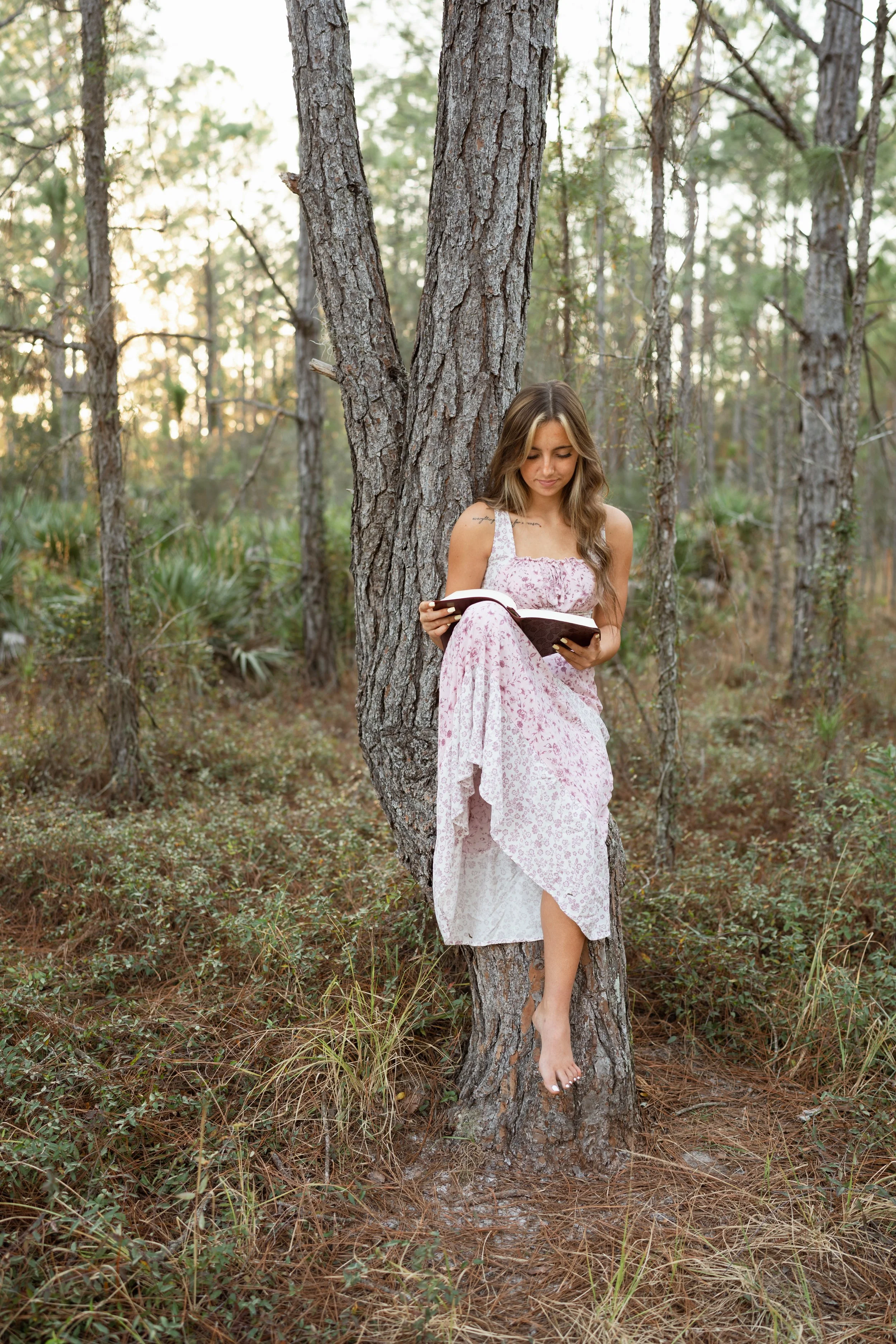 A young woman in a pink and white dress leaning against a tree in a forest, reading a book.