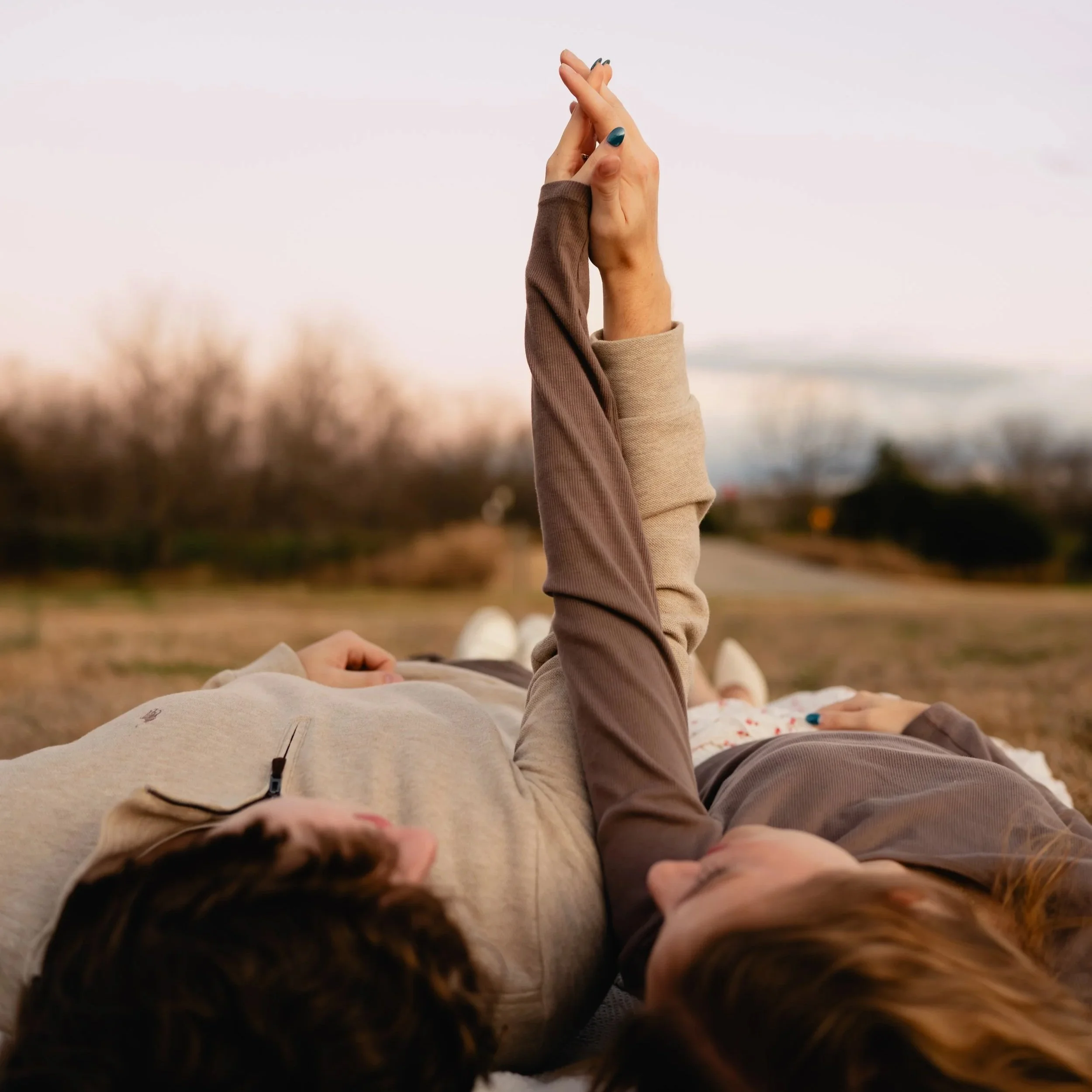 Two women lying on the grass outdoors, one with a brown long-sleeve shirt and the other with a light beige zip-up sweater, holding hands with fingers crossed, during sunset or late afternoon.