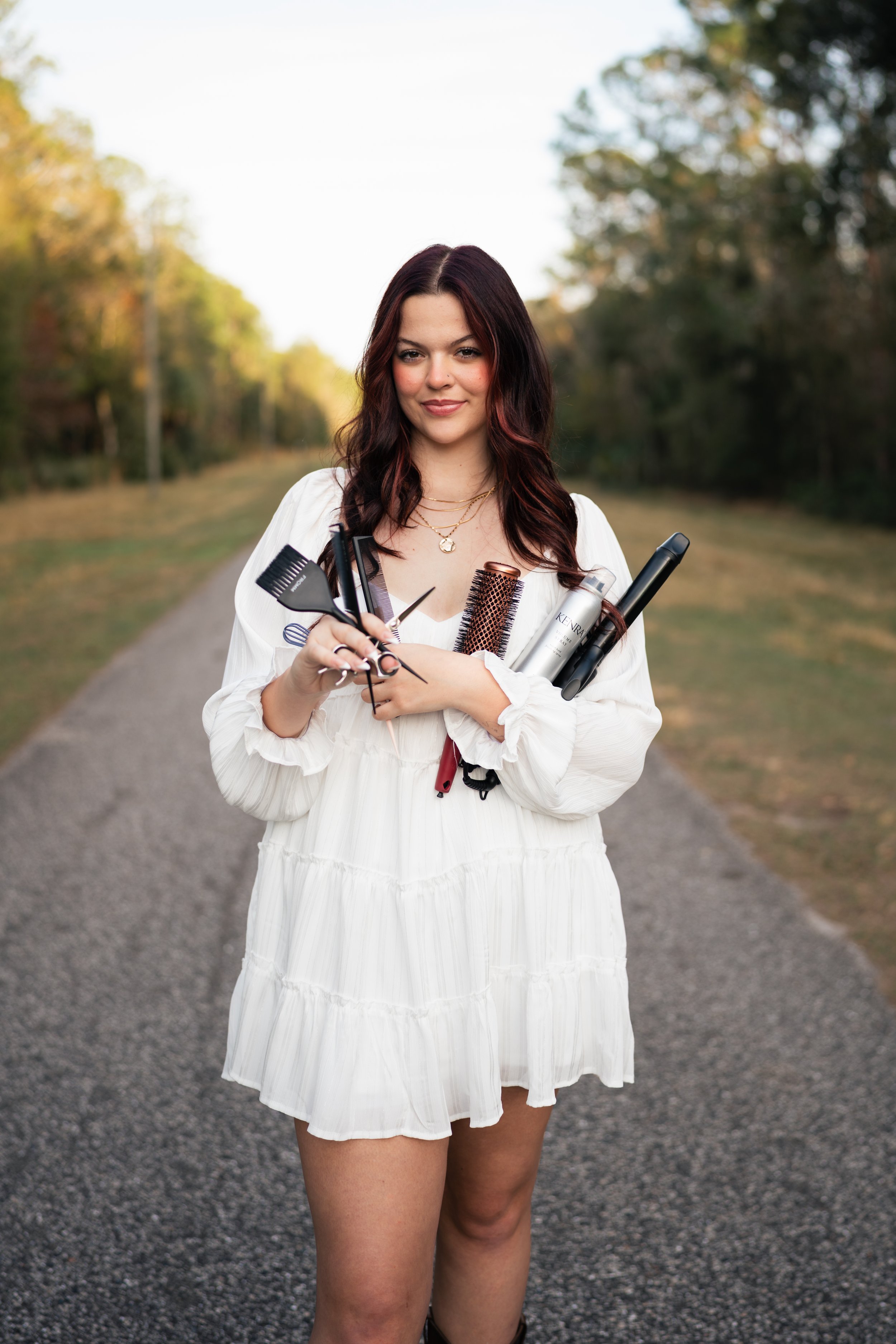 A woman standing on a gravel path outdoors in a white dress, holding various hair styling tools and products.