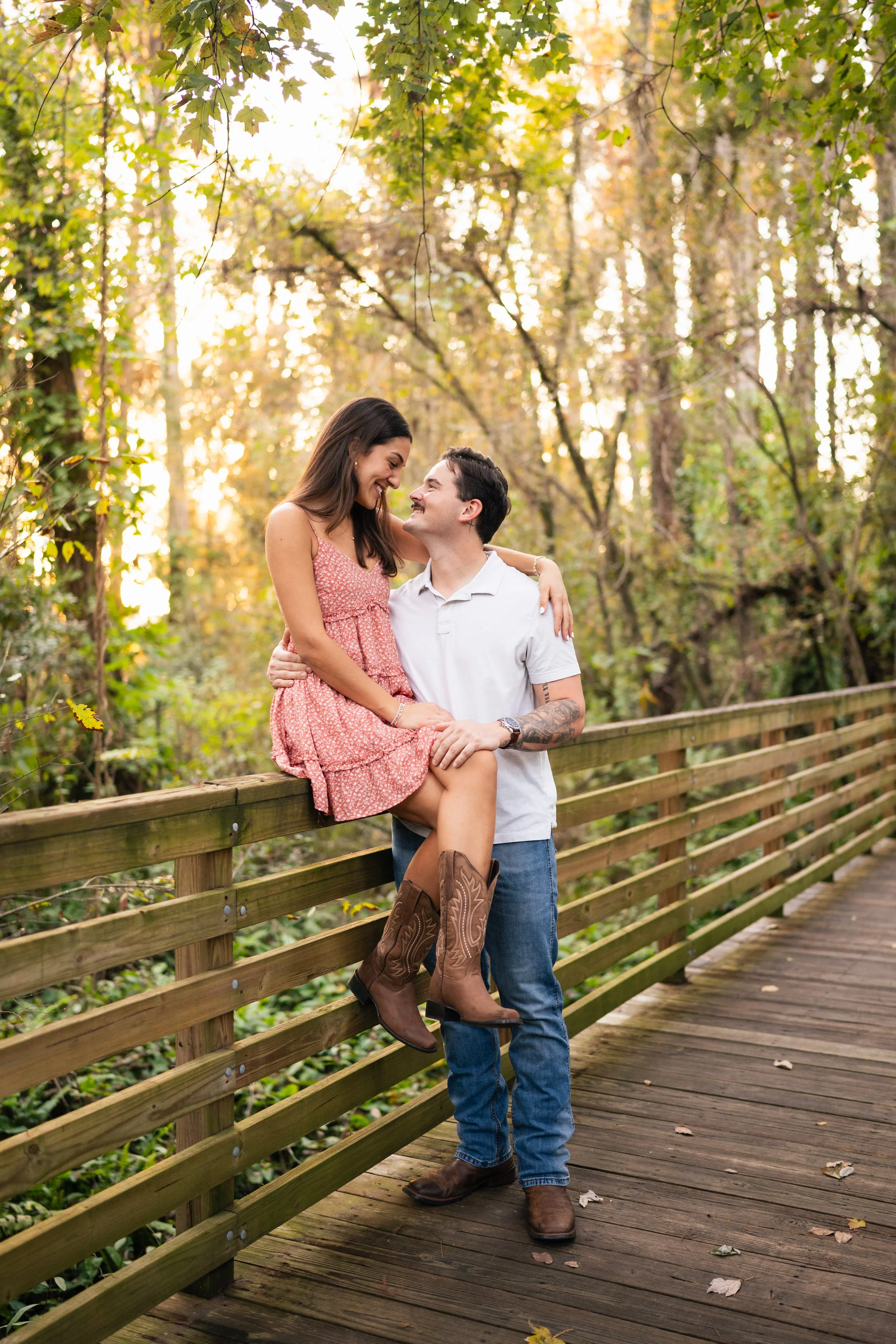 A smiling woman in a pink dress and brown cowboy boots sitting on a wooden railing, being held by a man in a white polo shirt and jeans on a tree-lined wooden bridge in a forest during autumn.