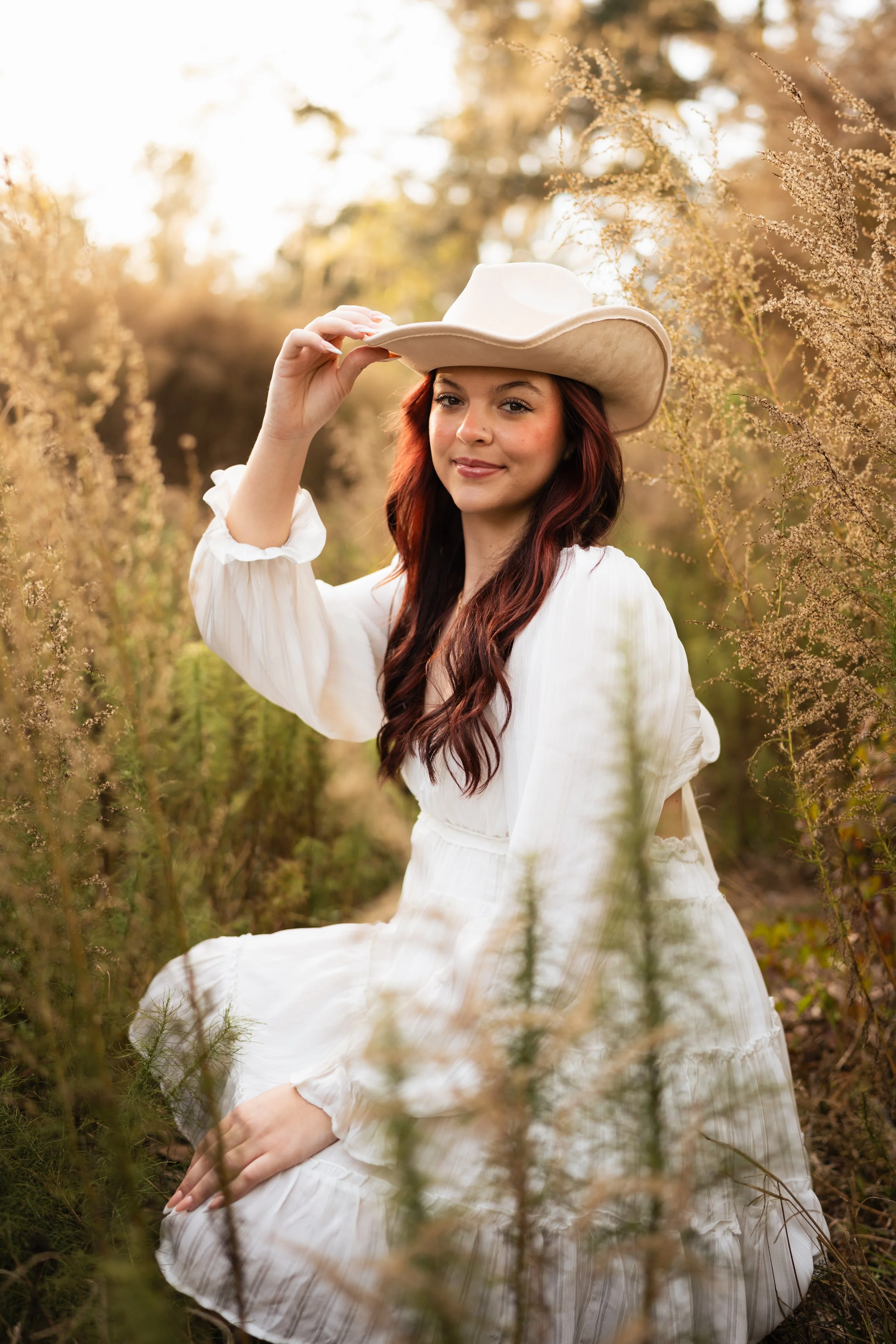 A young woman with long red hair wearing a white dress and cowboy hat, sitting amidst tall grasses and wildflowers in a natural outdoor setting during sunset.