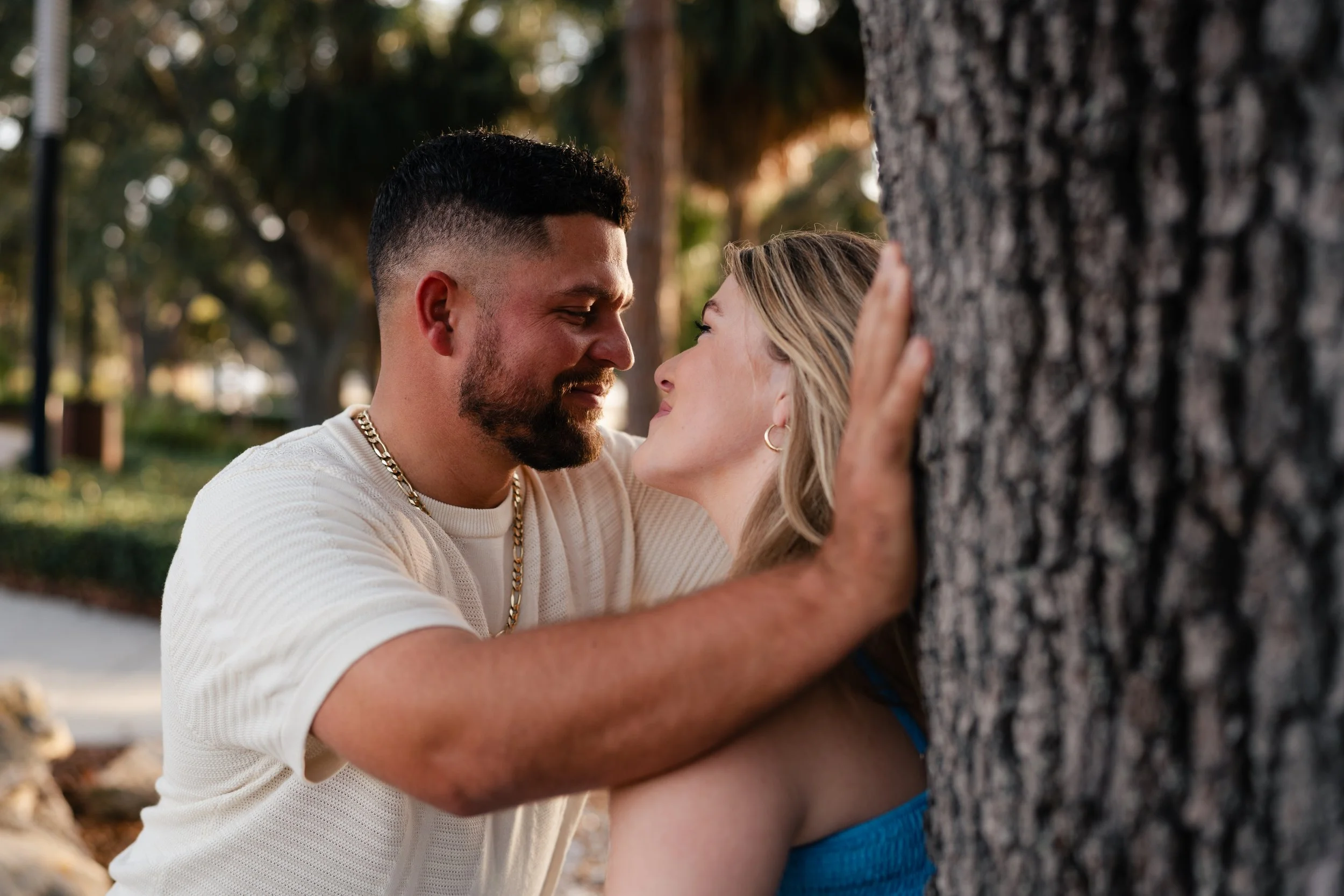 A couple is outside near a tree, about to kiss, with the man touching the tree and gazing into the woman's face.