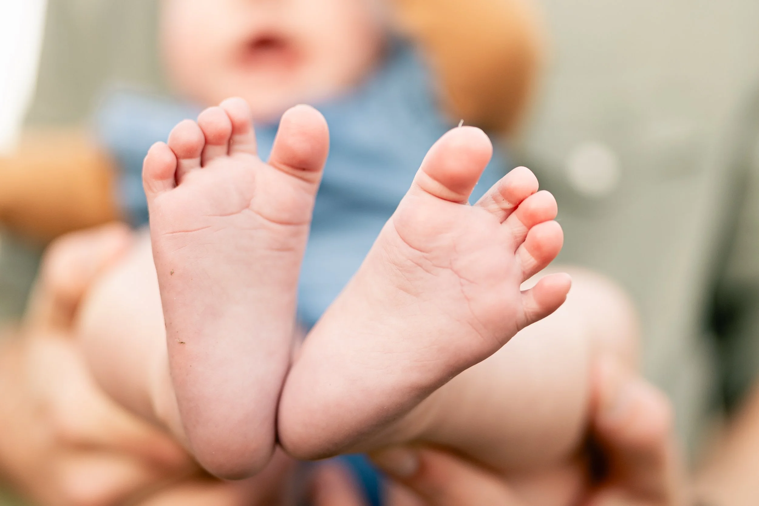 Close-up of a baby's feet with toes spread, held by an adult, with the baby's face slightly blurred in the background.