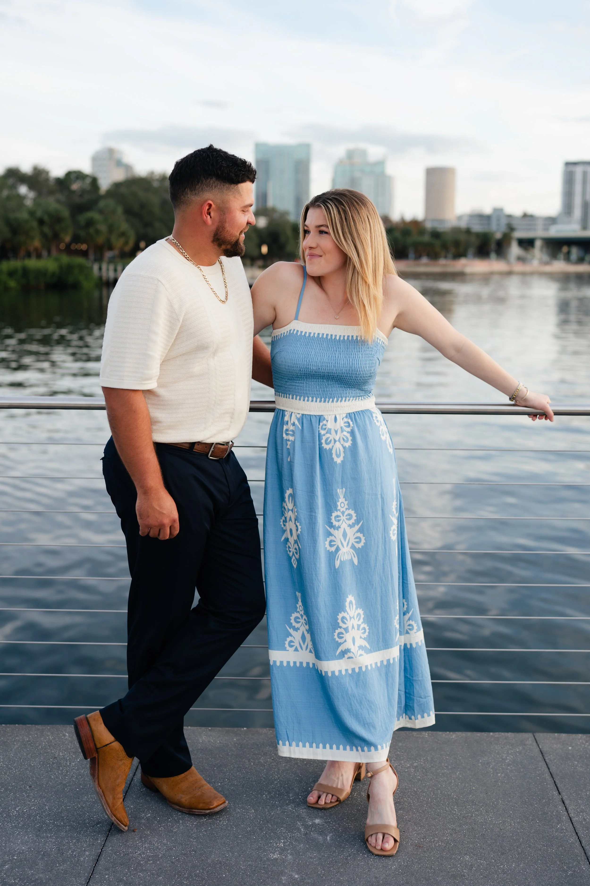 A man and woman standing close to each other on a dock by the water, with city buildings in the background, during daytime.