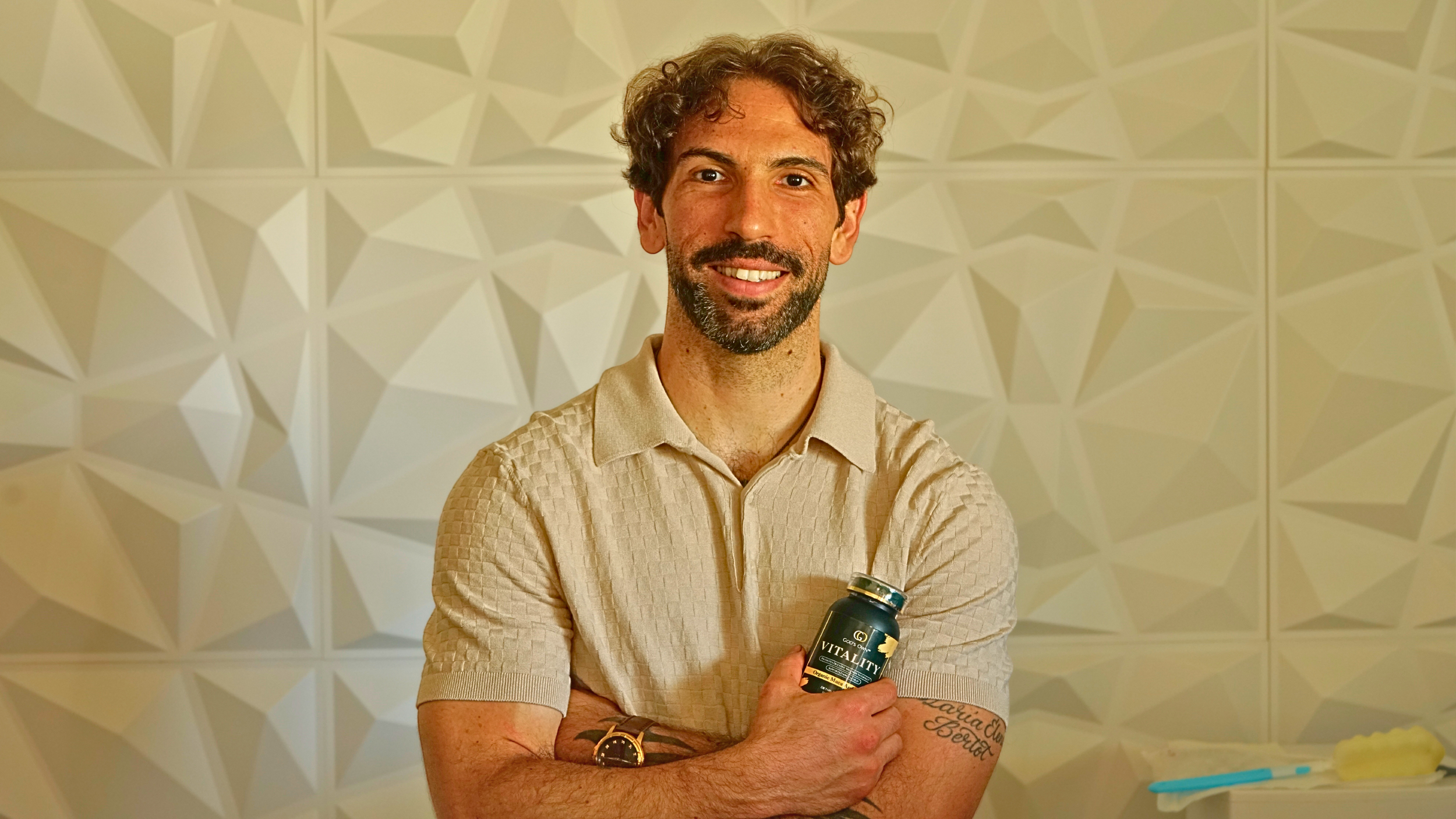 A man with curly brown hair and a beard smiling, holding a bottle labeled 'Vitality,' standing in front of a geometric patterned wall.