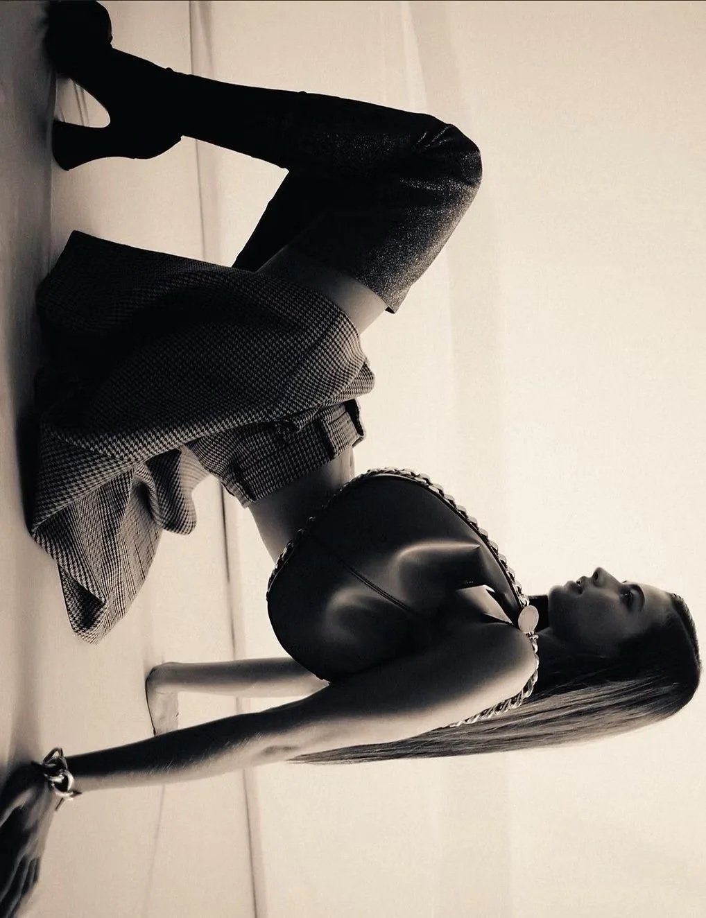 Black mannequin dressed in a sleeveless top and plaid skirt, reclining on the floor with one arm extended, in a minimalist indoor setting with plain walls.