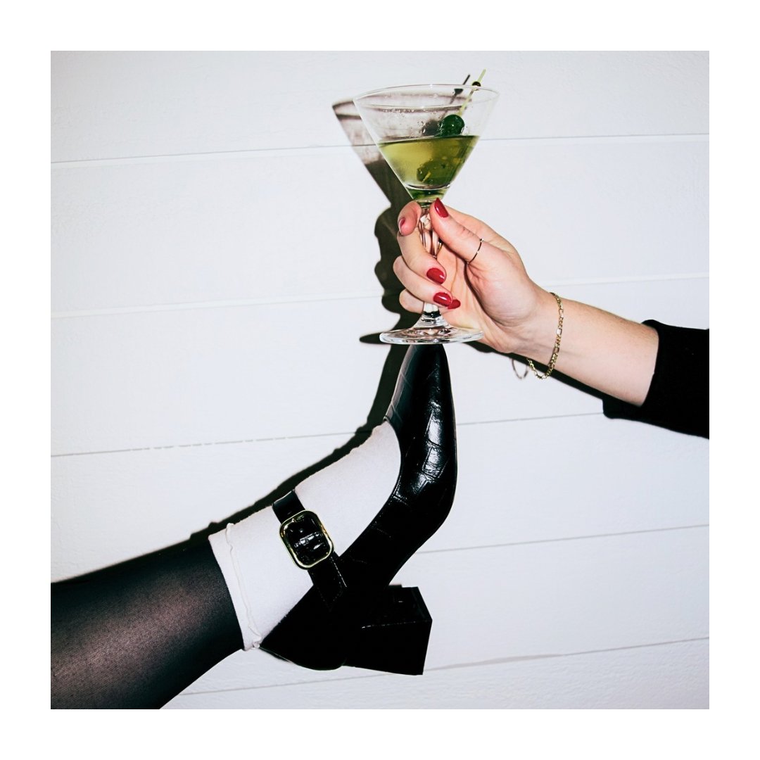 A woman with red painted nails and a gold bracelet holding a martini glass with green olives, balancing the glass on a high-heeled black shoe worn by a person with black tights and a white sock, against a white wall background.