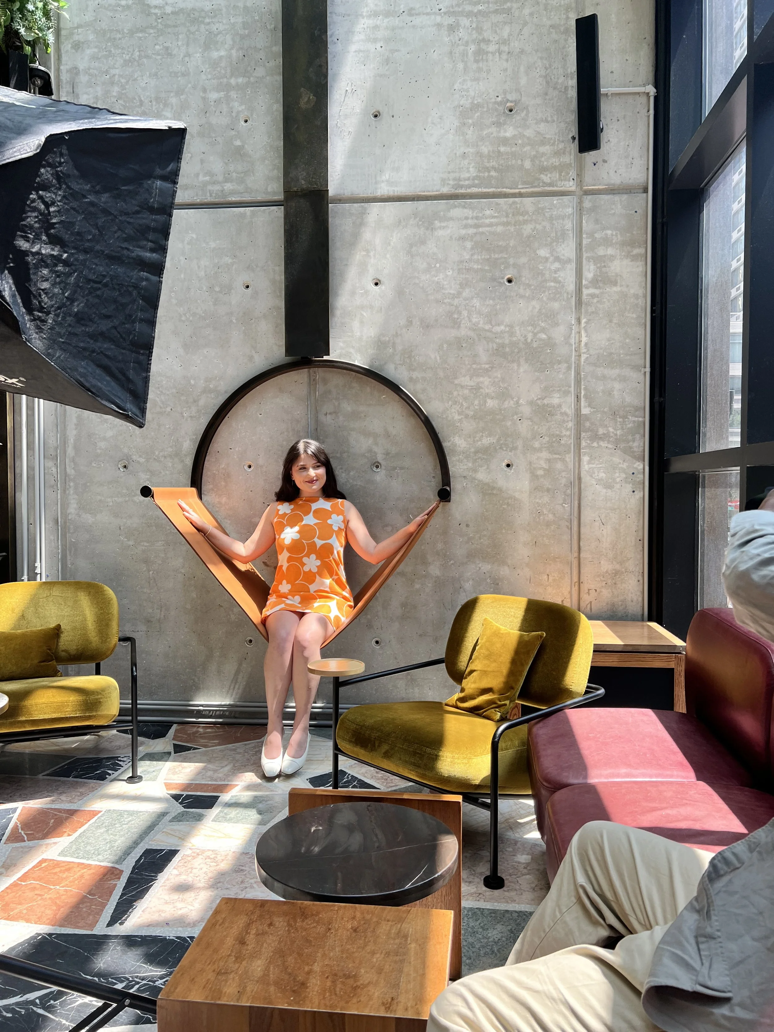 A woman in a sleeveless orange and white floral dress sits in what looks like a hammock chair inside a modern lounge area with concrete walls, surrounded by colorful chairs and tables, near large windows letting in natural light.