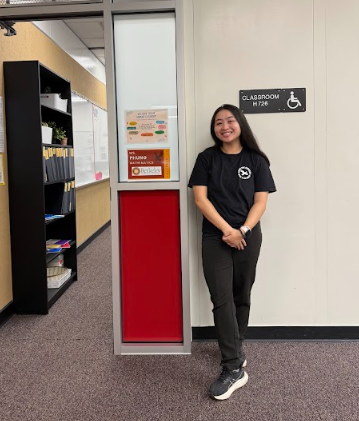 A smiling young woman standing outside a classroom marked for wheelchair accessibility in a school hallway.