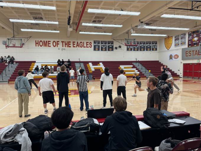 People playing basketball on an indoor court in a gymnasium. The walls have banners and a sign reading 'HOME OF THE EAGLES.'