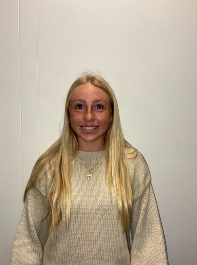 A young woman with light brown hair, wearing hoop earrings, a necklace with a heart-shaped pendant, and a dark green spaghetti strap top, standing against a plain off-white wall.