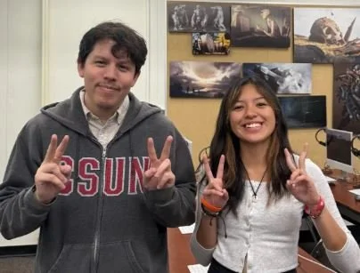 Two young people smiling and making peace signs in an indoor setting with framed pictures on the wall behind them.