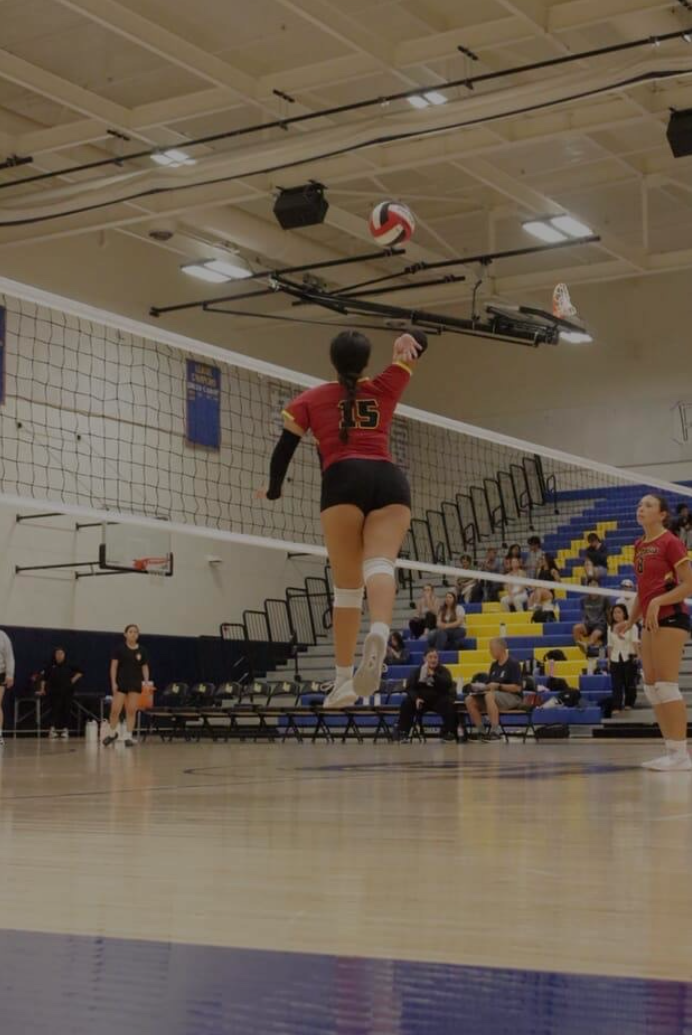A female volleyball player in mid-air preparing to spike the ball during a game in a gymnasium with spectators in the background.