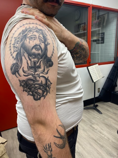 A man with a large tattoo of Jesus Christ's face with a crown of thorns on his upper arm, featuring a cross and a sacred heart. He has additional tattoos on his forearm and is wearing a white sleeveless shirt.