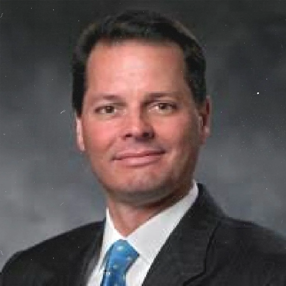 Photograph of a man in a black suit, white shirt, and blue tie, with dark hair, against a gray background.