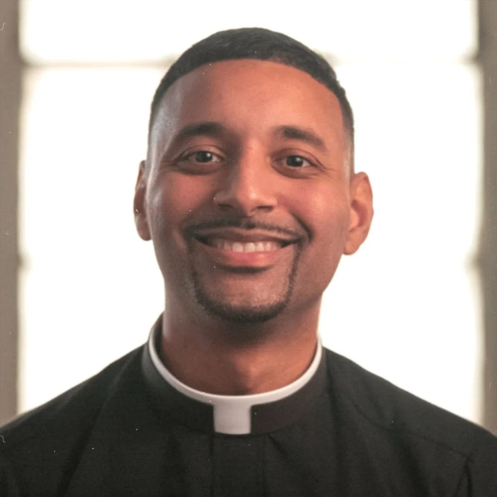 A smiling man dressed as a priest, wearing a black clerical shirt with a white collar, standing indoors with a bright window in the background.