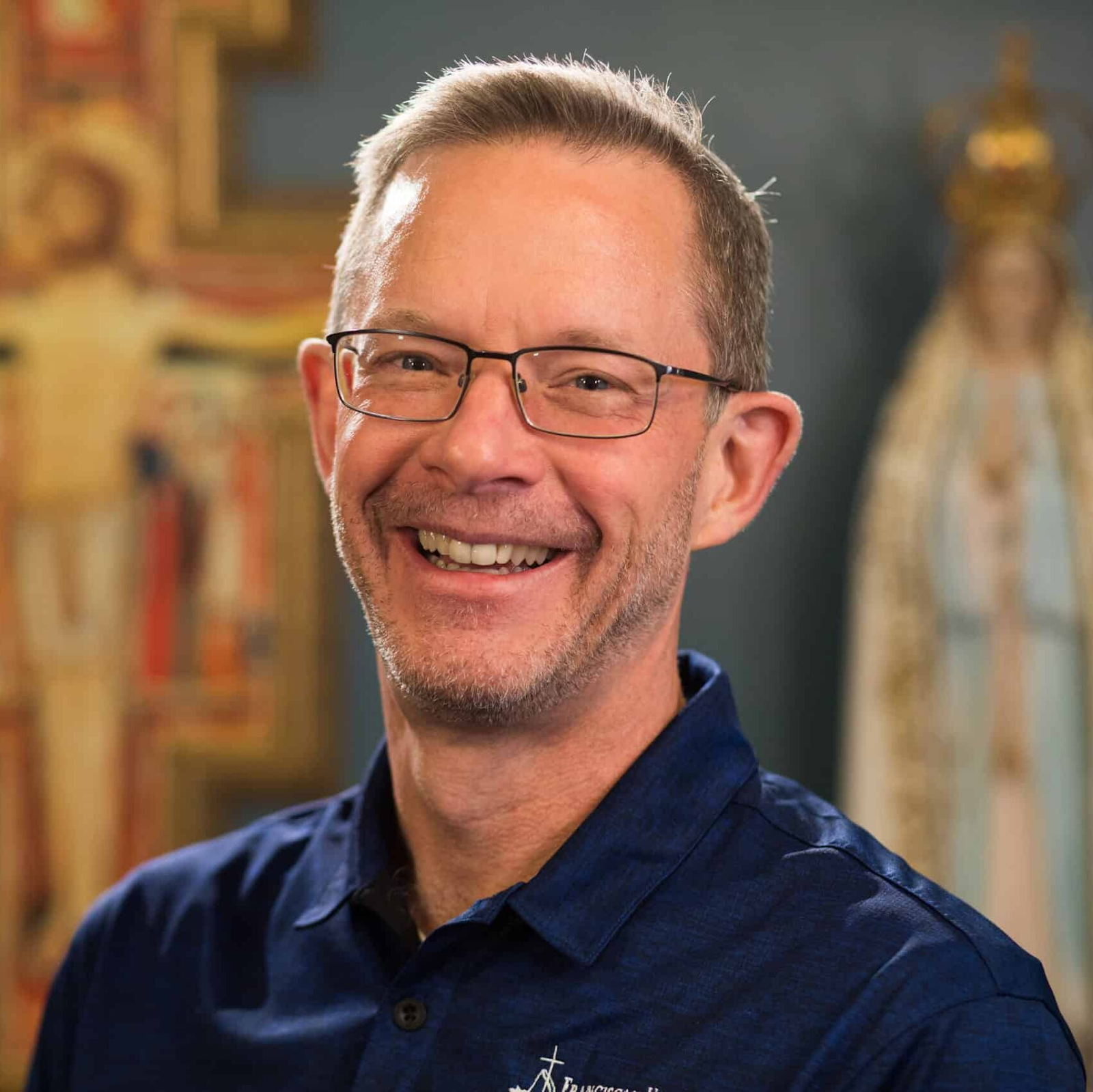 A smiling middle-aged man with glasses, short light brown hair, and facial hair wearing a dark blue shirt, indoors with religious artwork in the background.