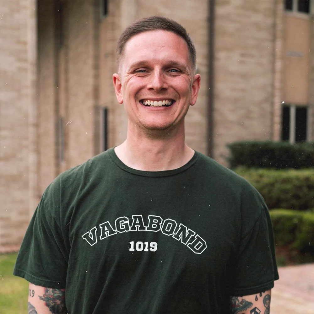 A smiling man with short brown hair, wearing a dark green T-shirt with the words 'VAGABOND 1019' printed on it, standing outdoors in front of a brick building and some greenery.