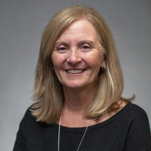 A smiling woman with shoulder-length blonde hair, wearing a black top and a necklace, against a plain gray background.