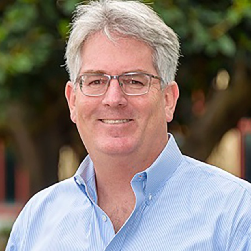 A middle-aged man with gray hair, glasses, and a blue button-down shirt, smiling outdoors with trees in the background.