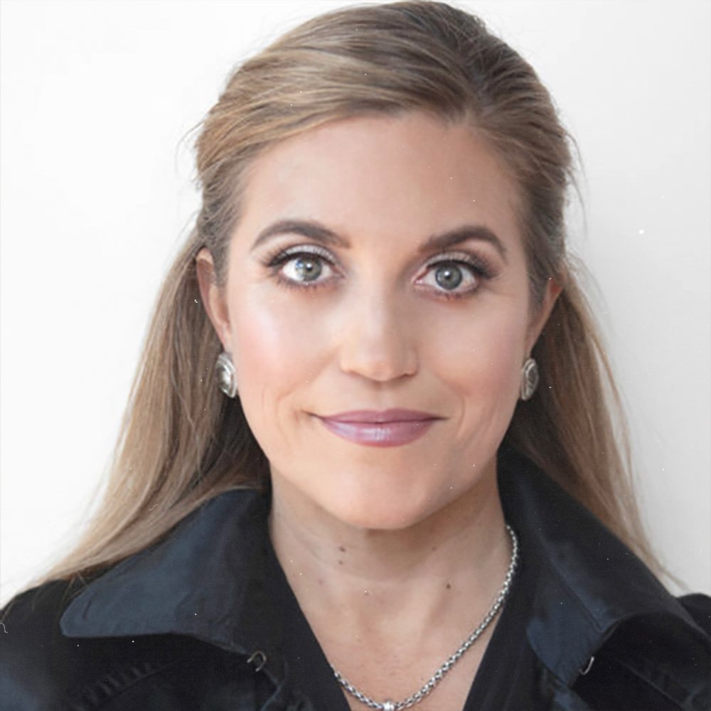 Close-up portrait of a woman with blue eyes, blonde hair, wearing earrings and a necklace, against a light background.