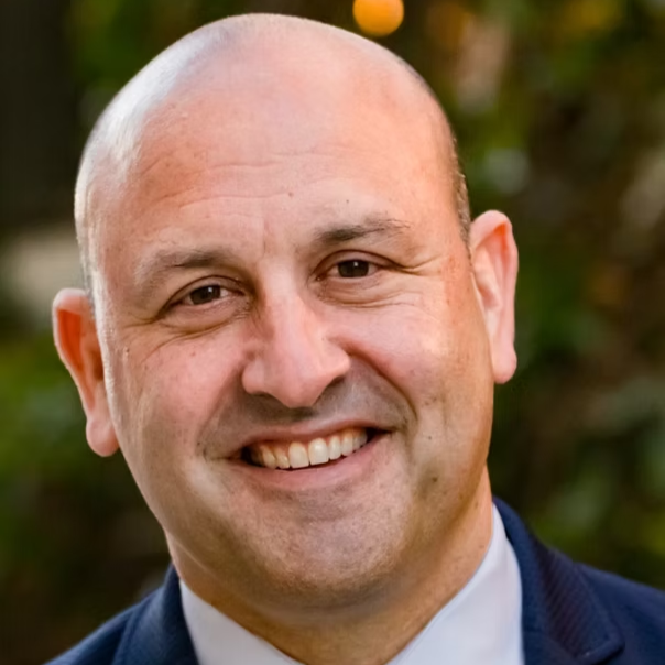 Close-up of a smiling man with a shaved head, wearing a dark suit and white shirt, outdoors with green and yellow blurred background.