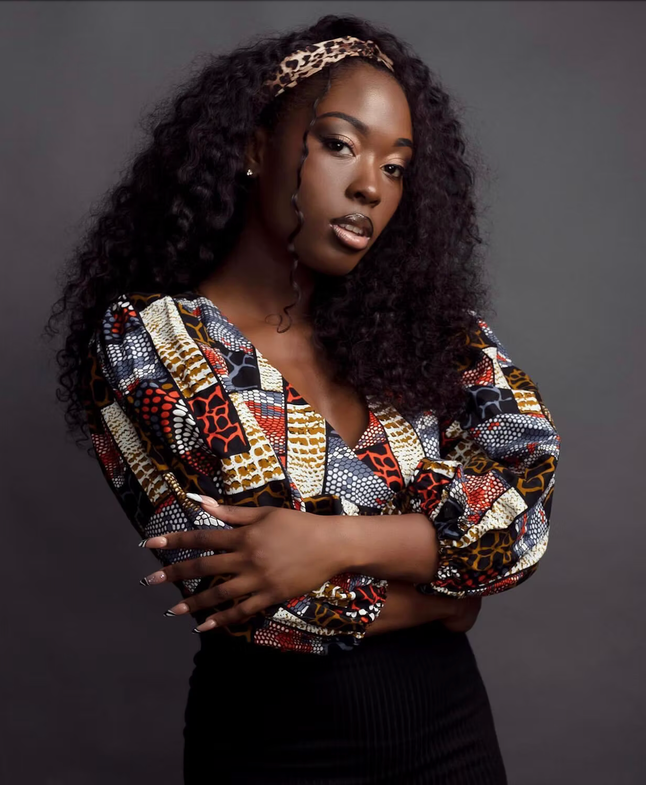 A woman with dark curly hair wearing a colorful patterned blouse with puffed sleeves and a leopard print headband, posing with her arms crossed against a gray background.
