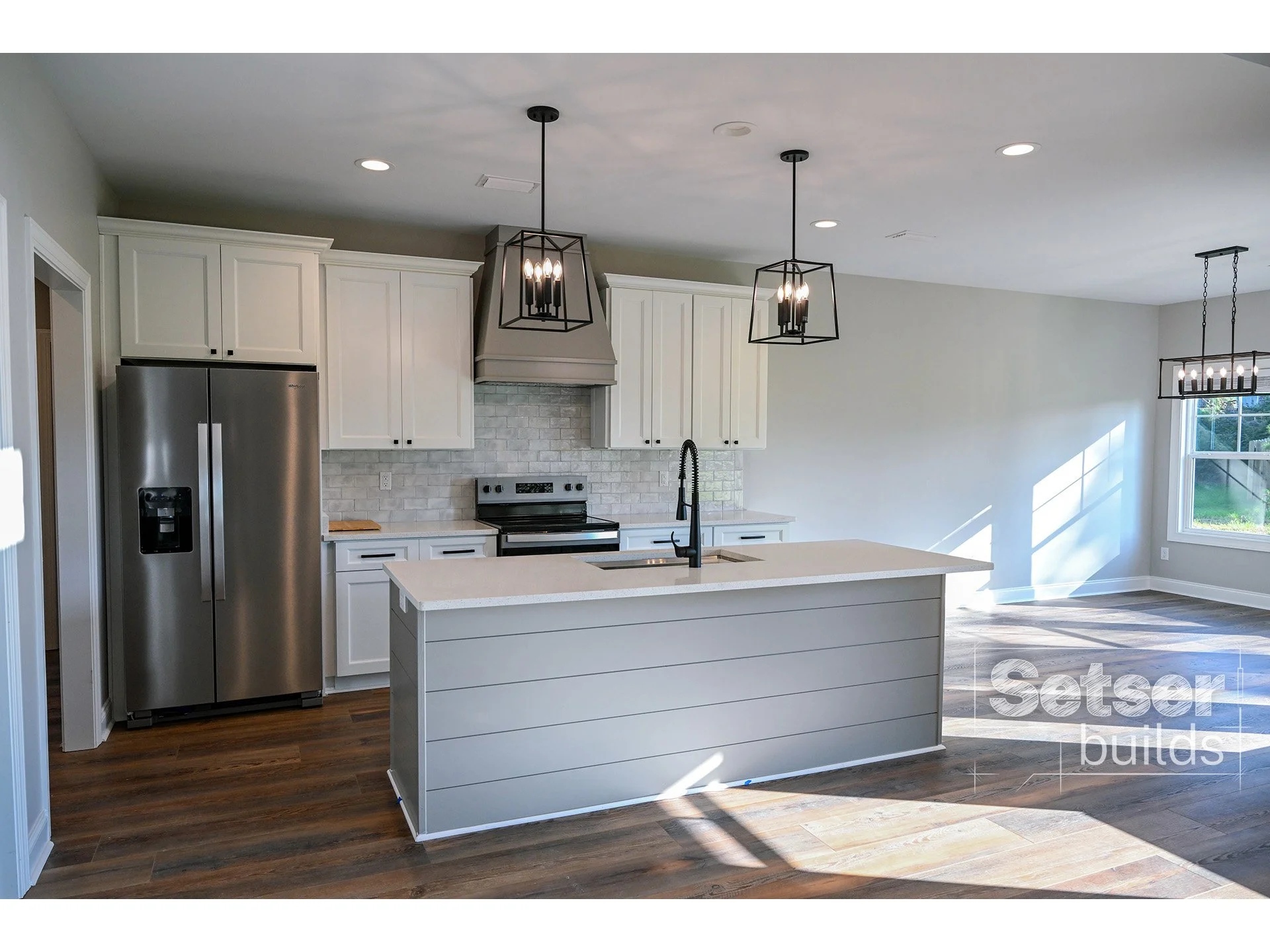 Modern kitchen with white cabinets, stainless steel refrigerator, island with black sink and faucet, brick backsplash, black pendant lights, hardwood floors, and a dining area with a chandelier near a large window.