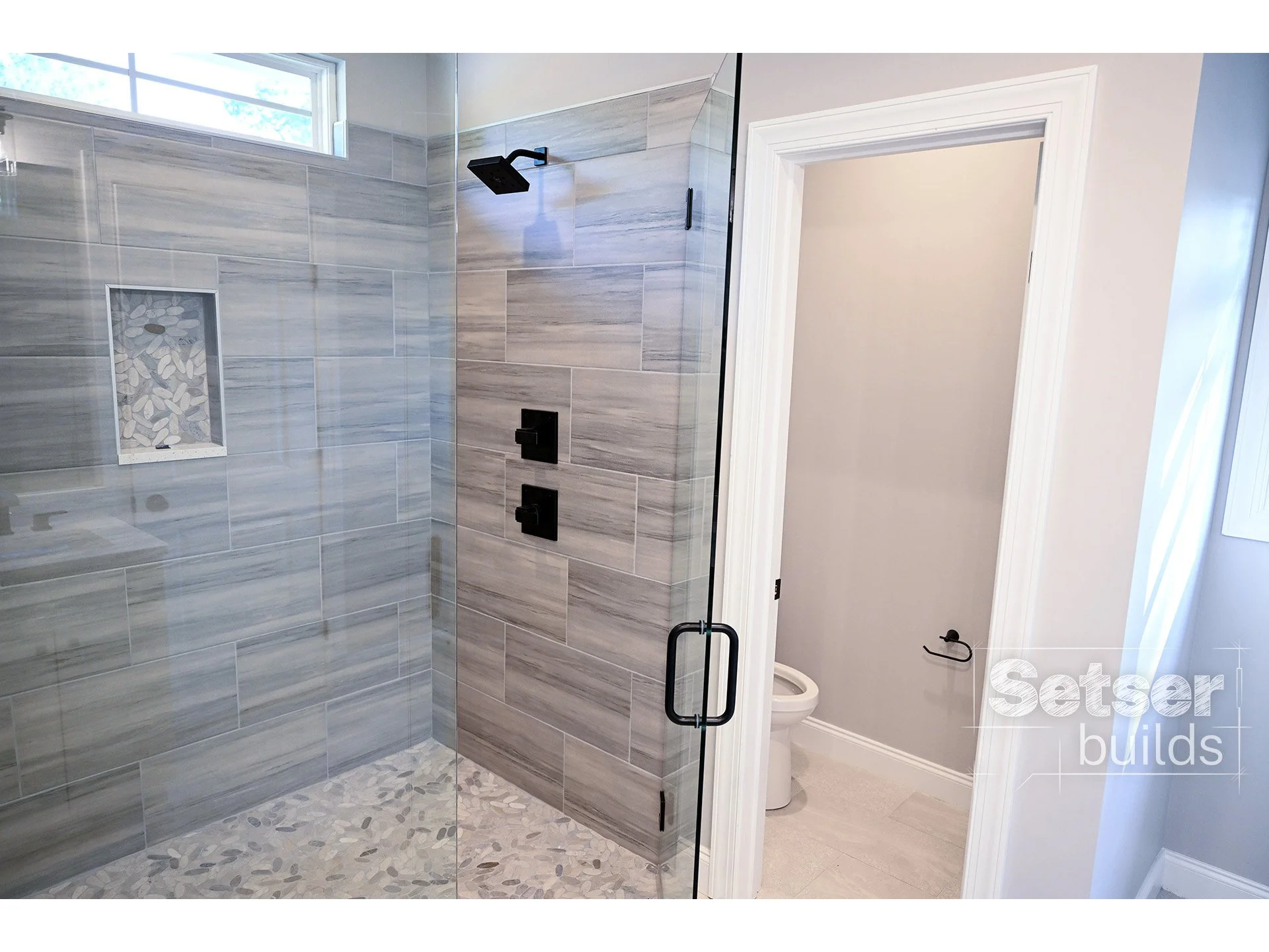 Modern bathroom with a walk-in shower featuring gray tiles, black fixtures, and a pebble floor. A window near the ceiling allows natural light, and a door leads to a small toilet room.