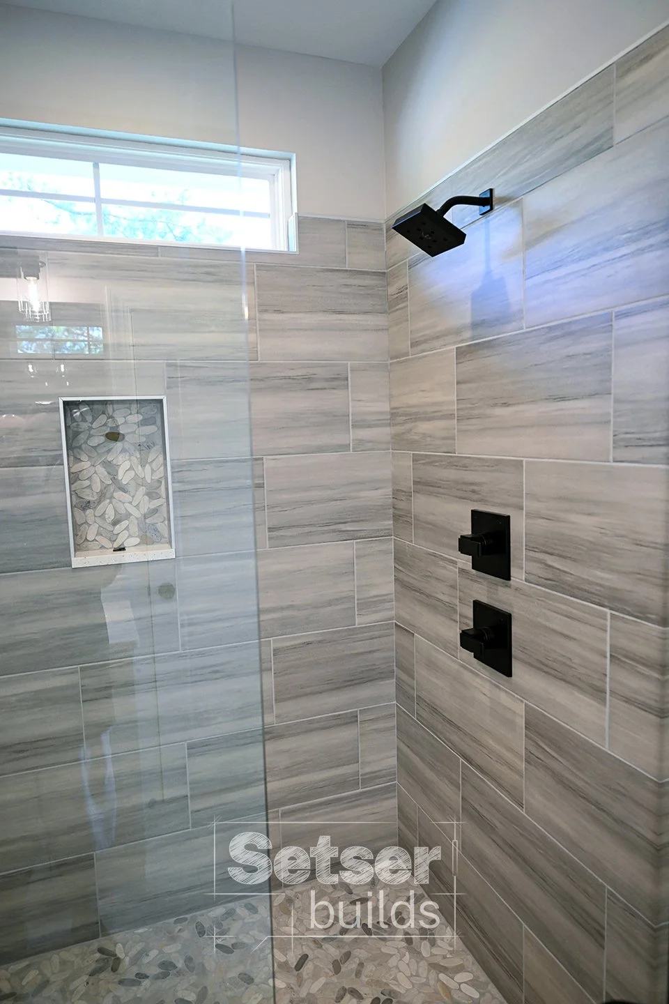 A modern walk-in shower with grey marble wall tiles and pebble-patterned shower floor. There is a recessed niche with pebble design on the wall and black shower fixtures, including a rainfall showerhead and two control handles. A window lets in natur