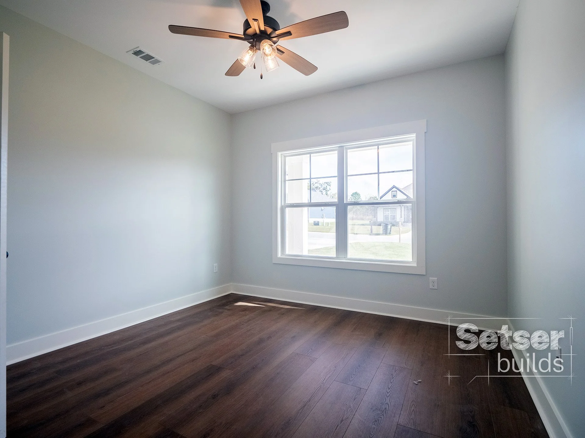 Empty room with light-colored walls, large window, dark hardwood floor, ceiling fan with lights.