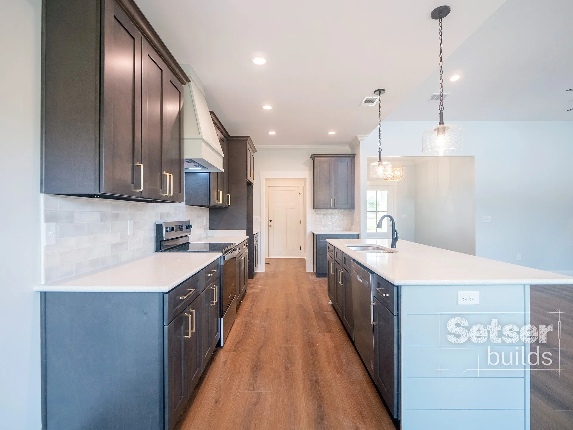 Modern kitchen with dark wood cabinets, white countertops, and a central island with a sink, pendant lights, and a wooden floor.