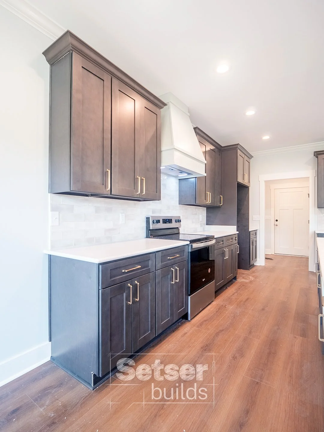 Modern kitchen with dark wooden cabinets, white countertops, a white range hood, stainless steel stove, white backsplash, and hardwood flooring.