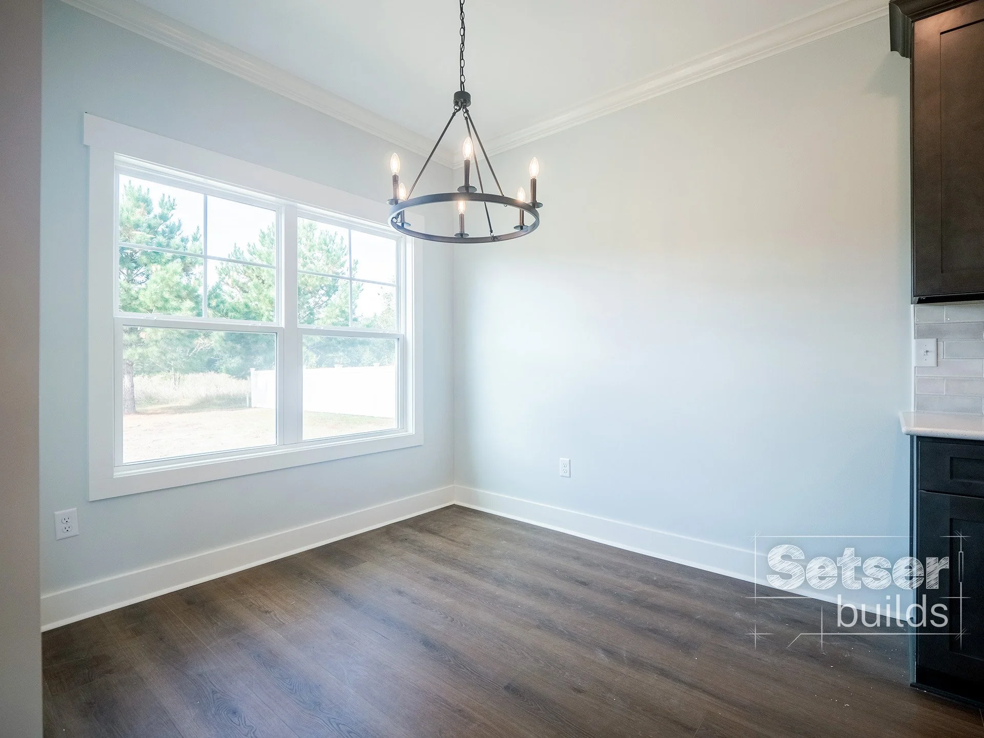 Empty dining area with large window, wooden floor, black chandelier, and part of kitchen cabinets visible.