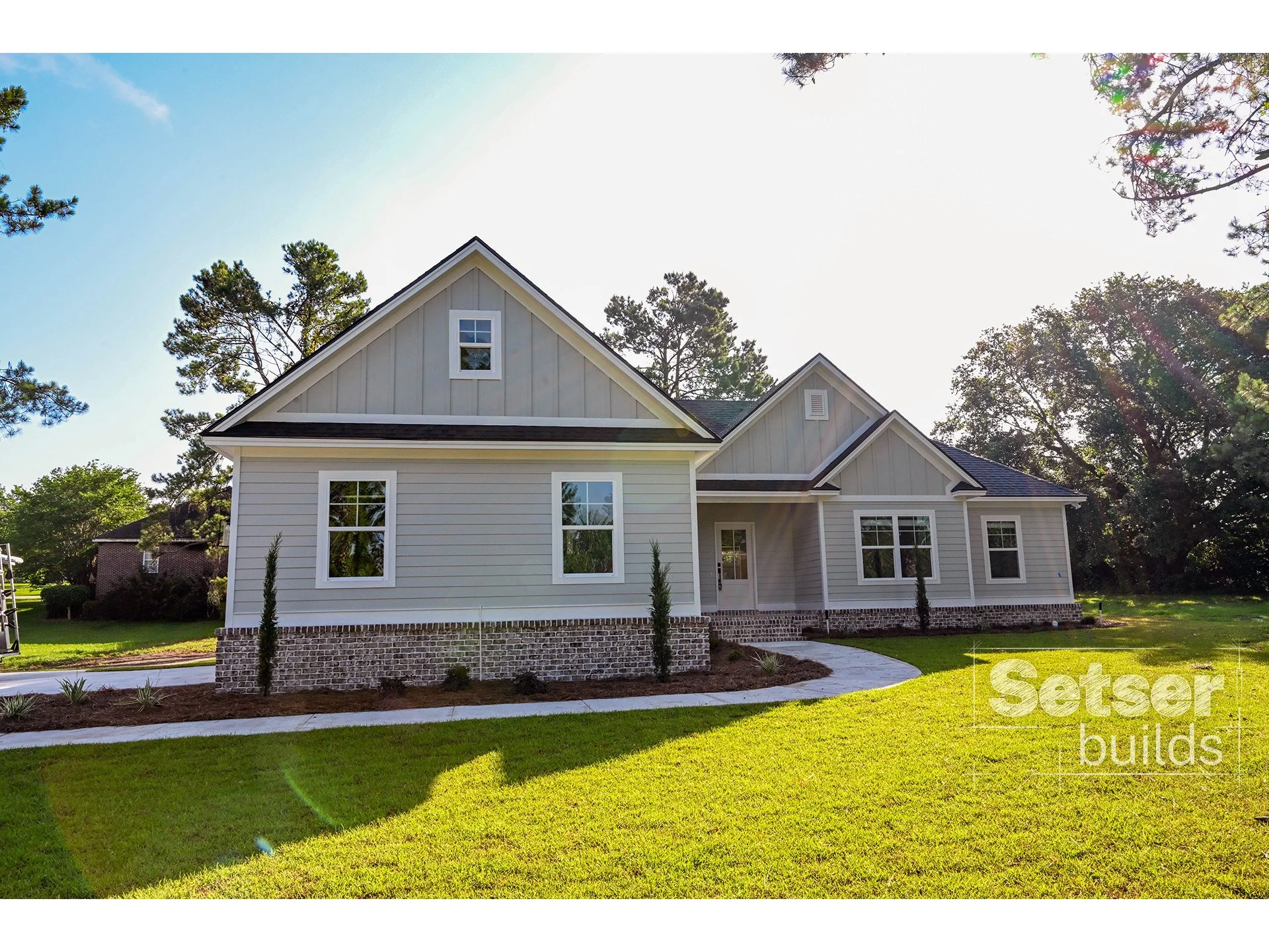 A new modern house with white siding and brick foundation, surrounded by a green lawn and trees, with a curved sidewalk leading to the front door.