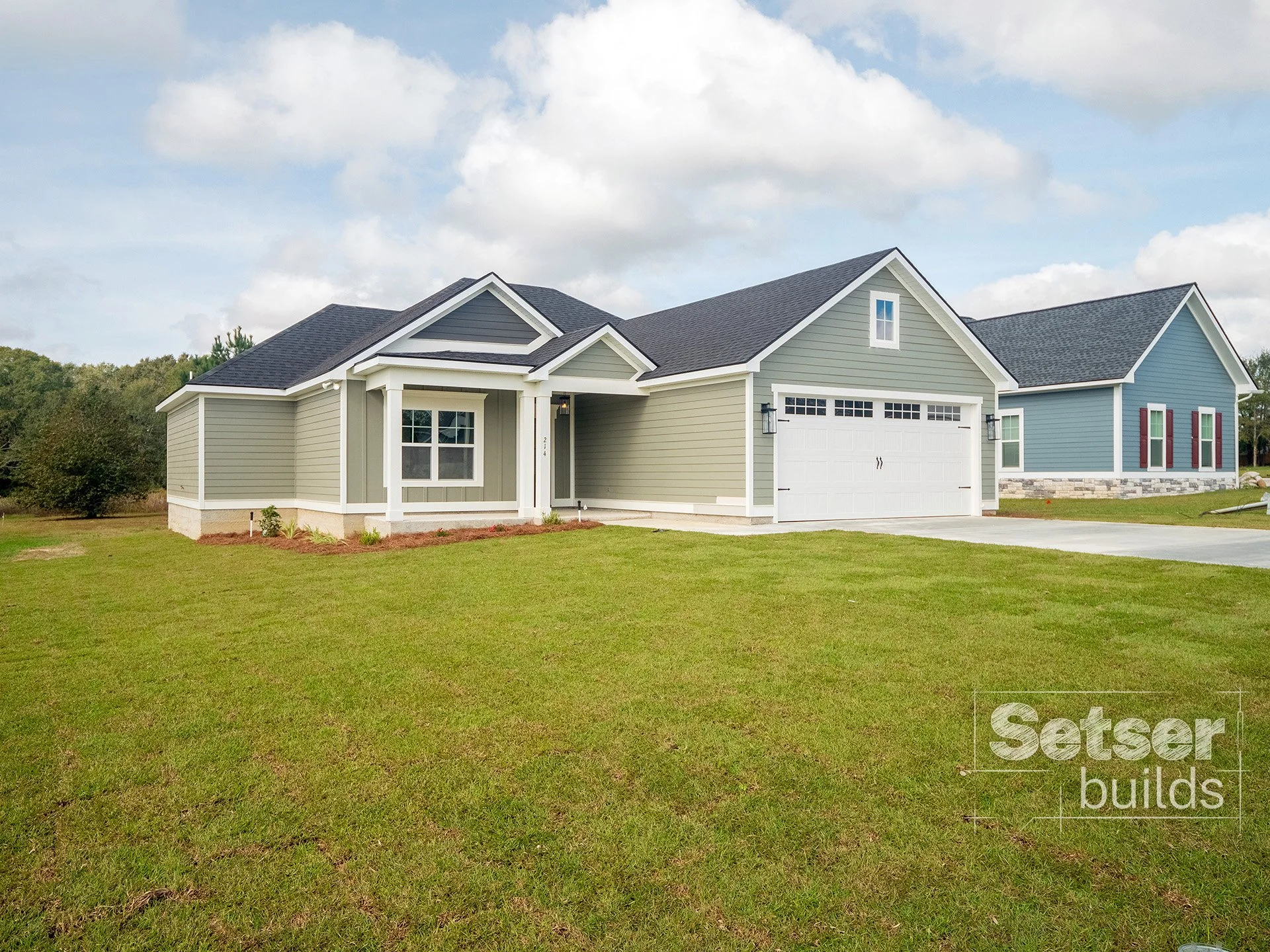 Front view of a new, single-story house with green and blue siding, white trim, black roof, and attached two-car garage, on a green lawn under a partly cloudy sky.