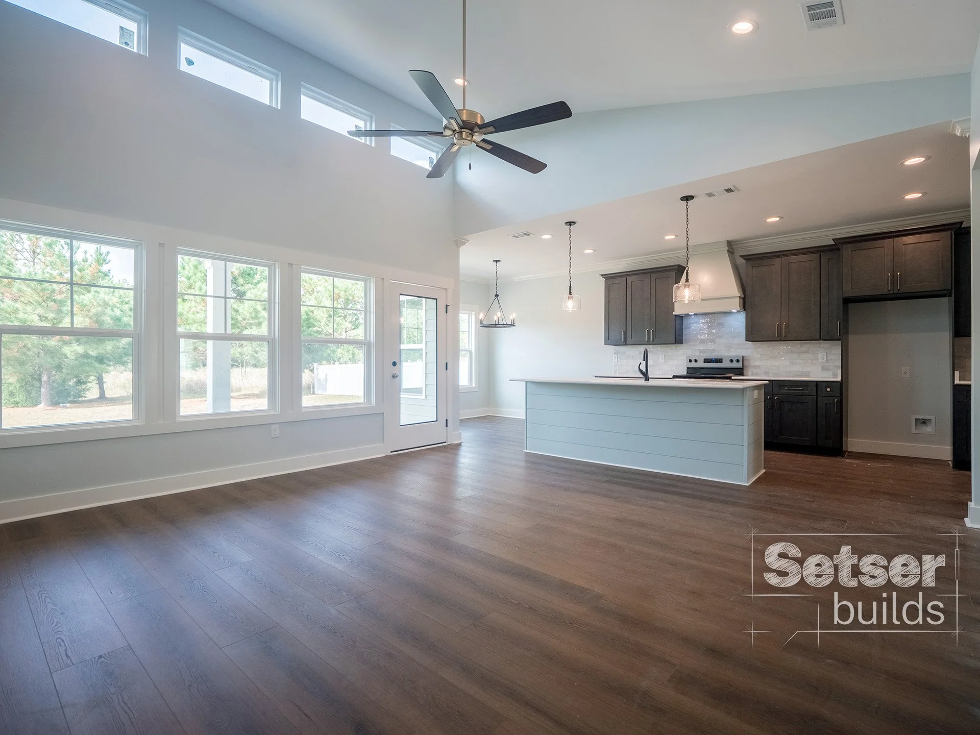 Open-concept living room and kitchen with large windows, wood flooring, a ceiling fan, and modern kitchen cabinets and appliances.