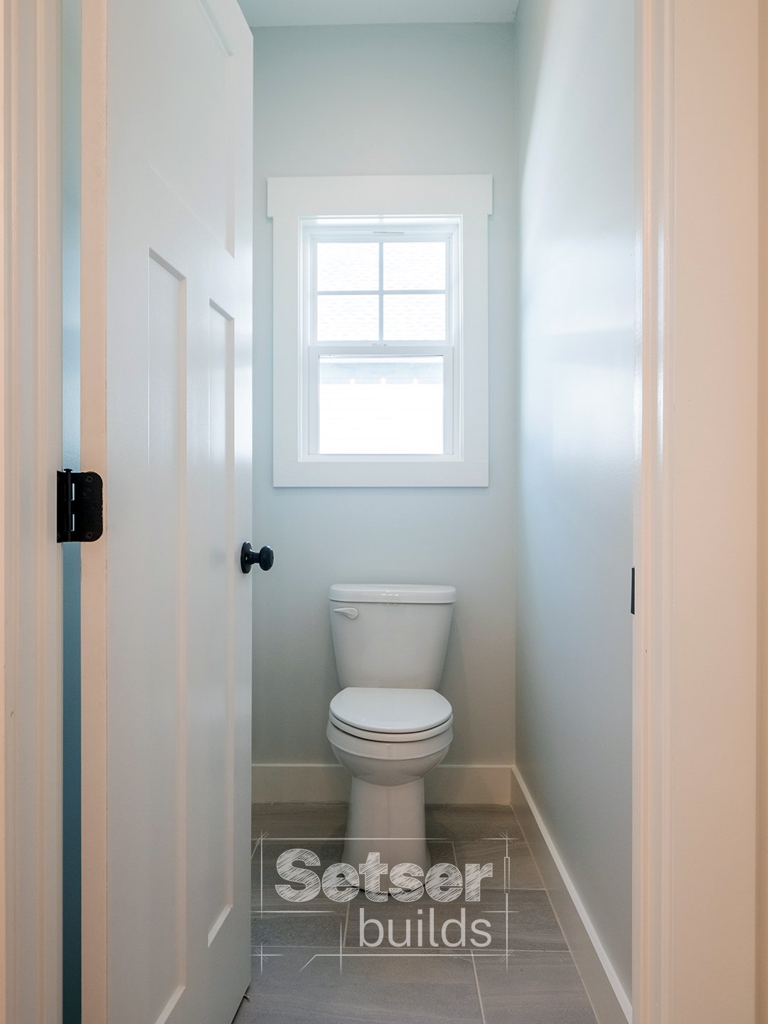 Small bathroom with a white toilet and a window above it, light-colored walls, and a wooden floor.