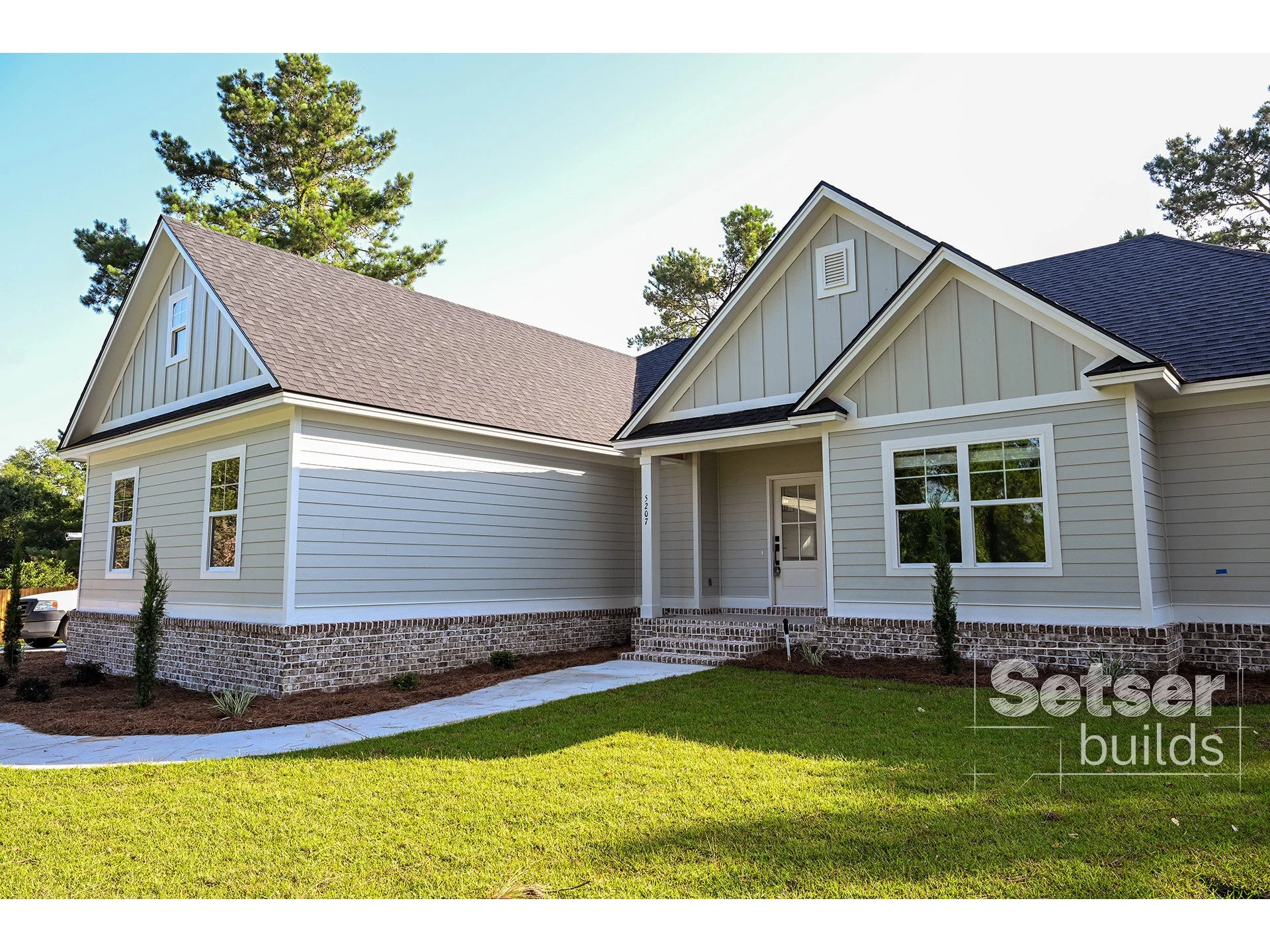 New house with beige siding, brick foundation, and brown shingle roof surrounded by green lawn and trees.