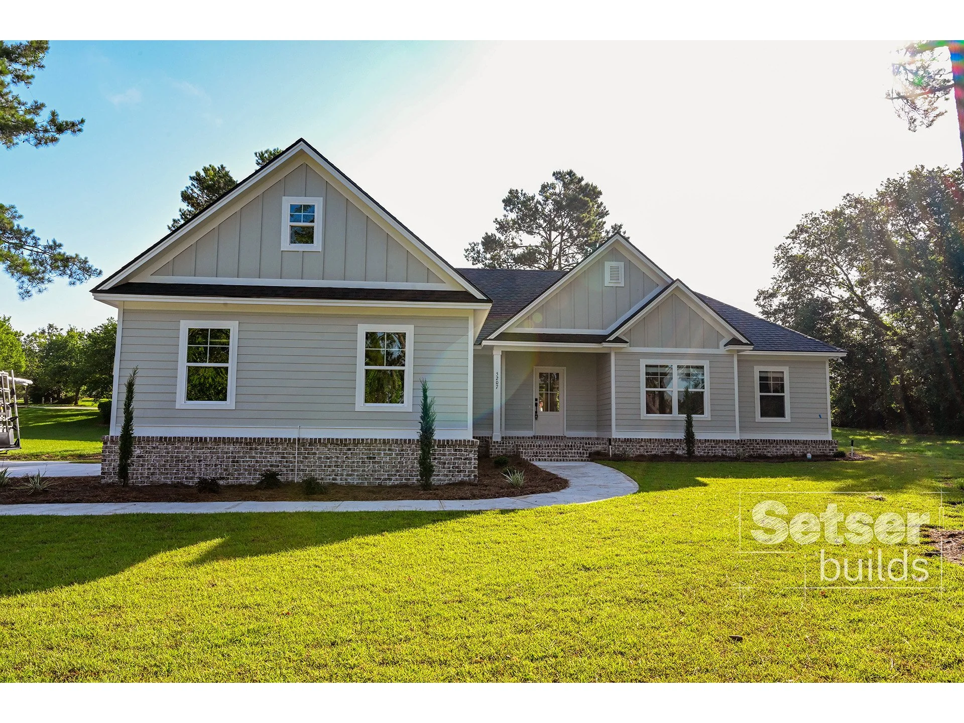 Newly built white house with vinyl siding and brick foundation, surrounded by a green lawn and trees, under clear blue sky.