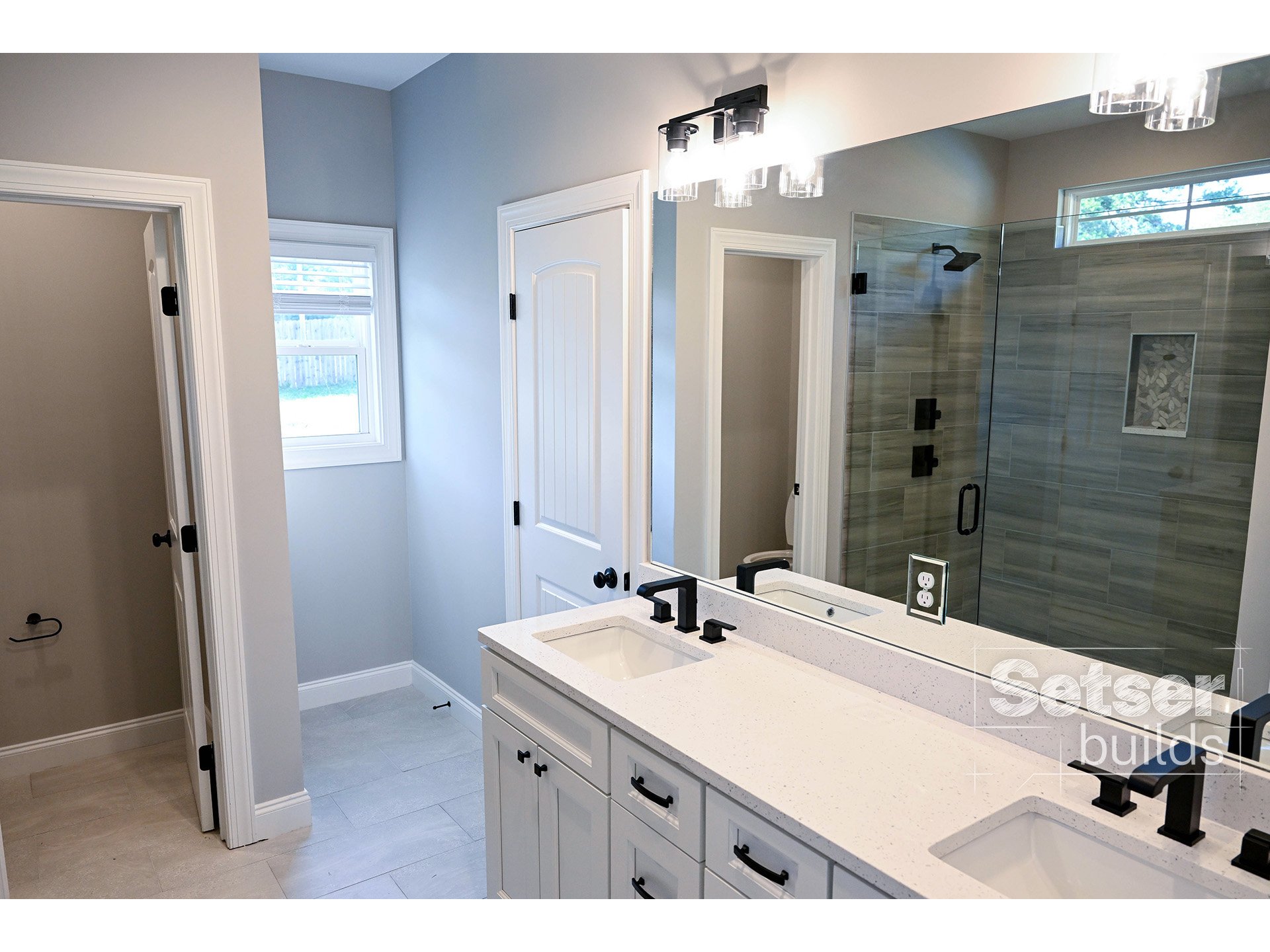 Modern bathroom with double vanity, black fixtures, large mirror, walk-in shower with glass door and gray tiles, small window, and beige tiled flooring.
