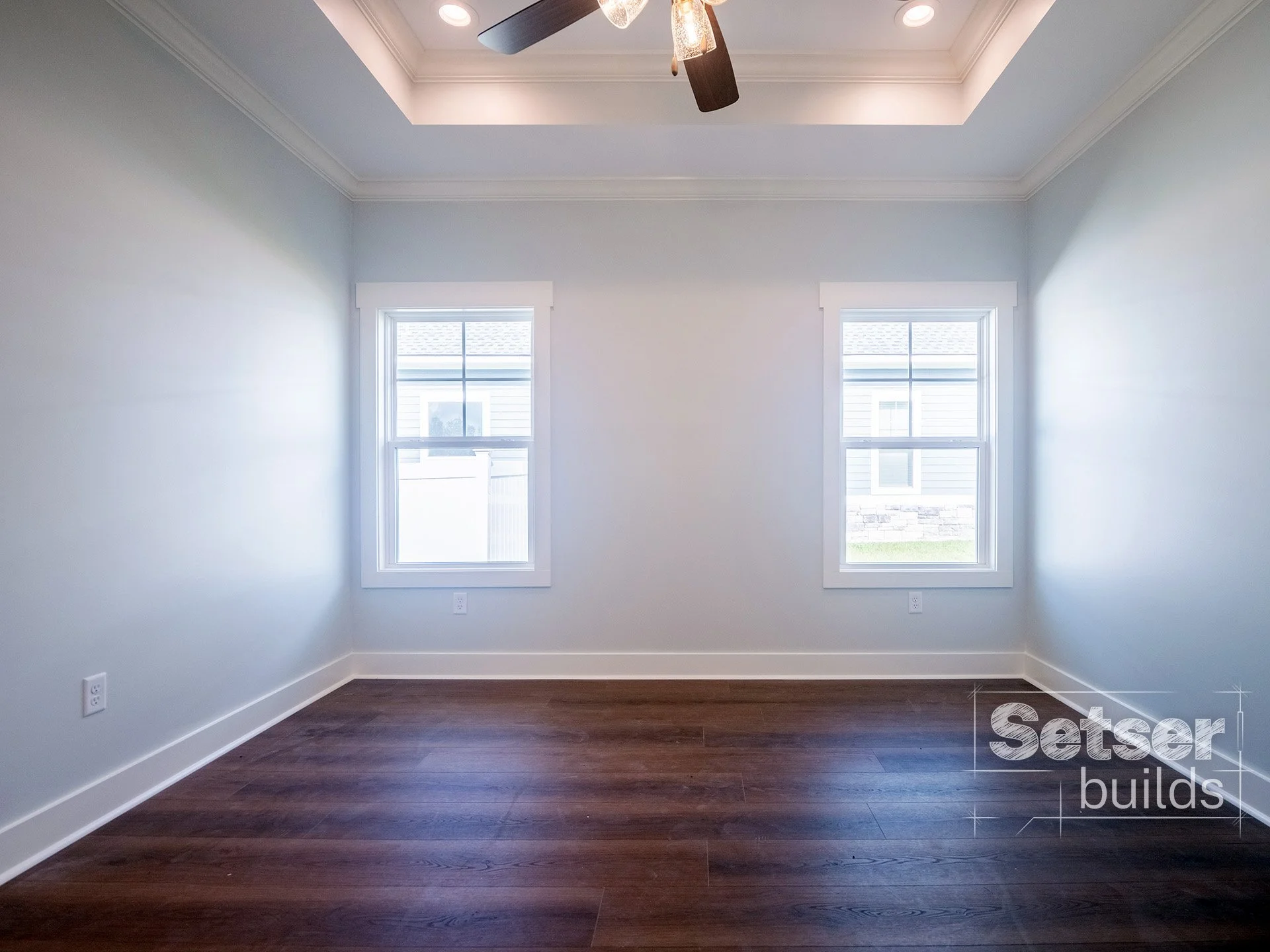 Empty room with two windows, white walls, a ceiling fan with light, and hardwood flooring.