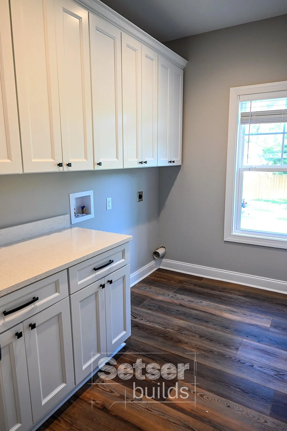 Empty laundry room with white cabinets, a window with blinds, hardwood floors, and a cable outlet with a pipe on the wall.
