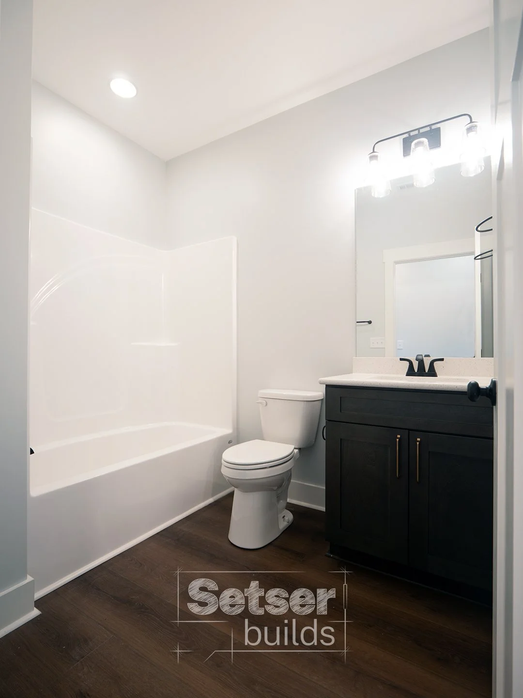 Bathroom with white bathtub, toilet, dark vanity with a sink, large mirror, and overhead light fixtures. Wooden floor. Minimalist design.