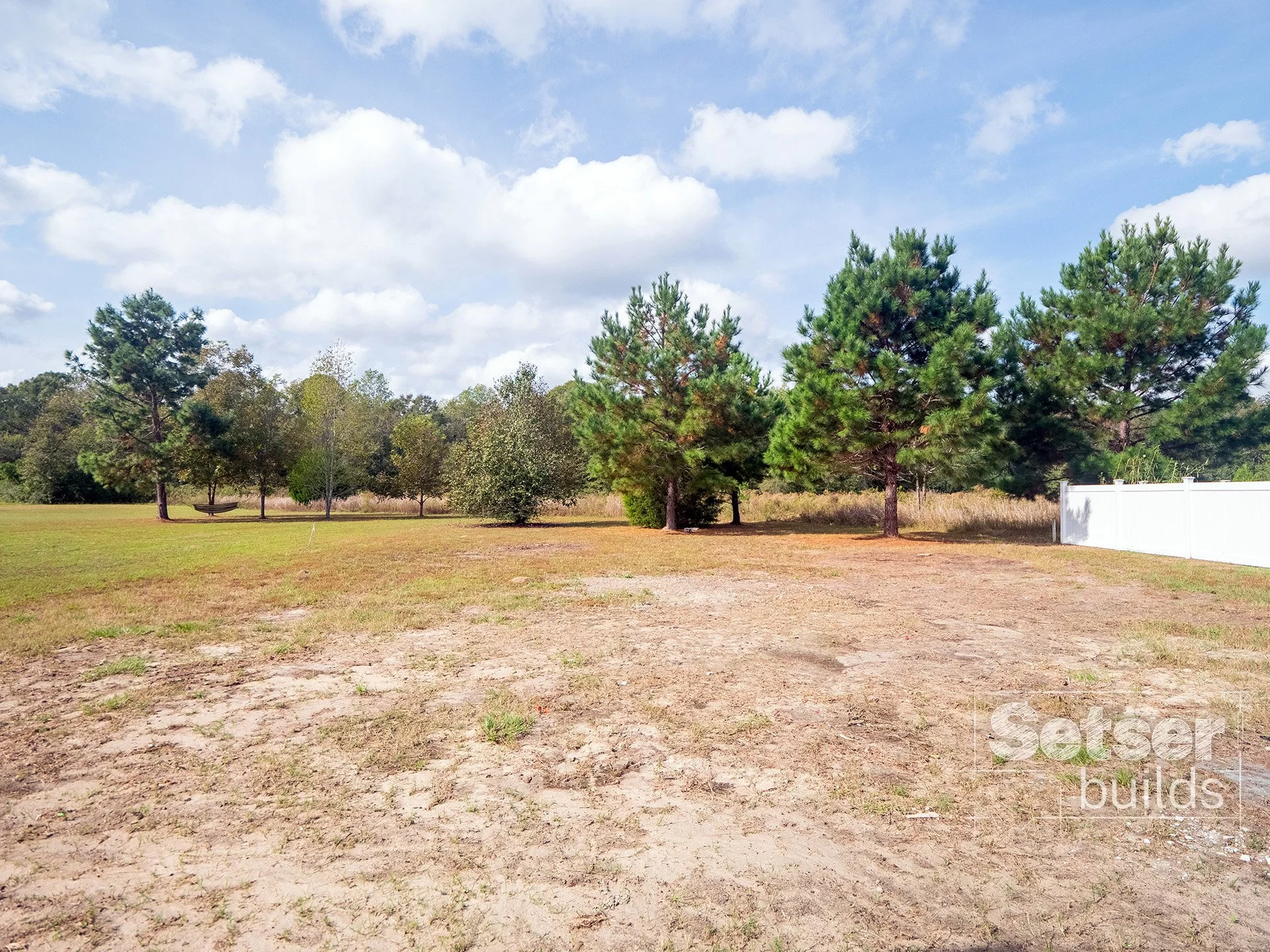 Open yard with a few trees, some greenery, and a white fence on the right side, under a partly cloudy sky.