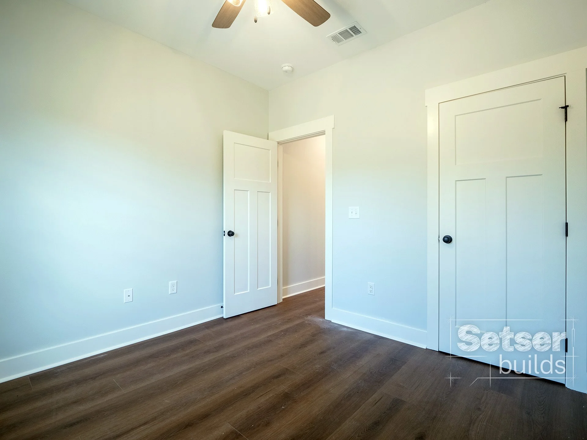 Empty room with white walls, a ceiling fan, dark wood floors, and two closed white doors, one with black knobs. The room has electrical outlets and a window not visible in the image.