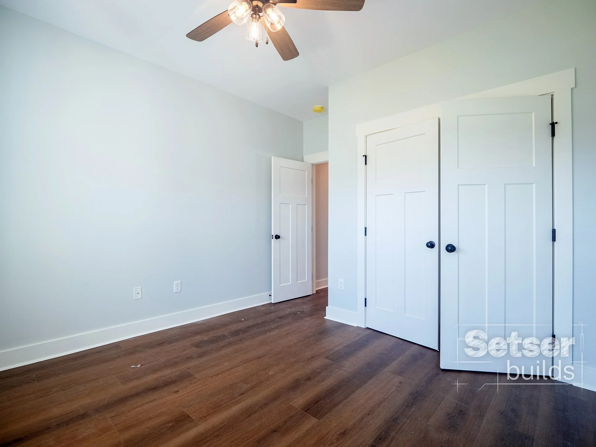 Empty room with white walls, dark wood flooring, a ceiling fan with five blades and four exposed bulbs, an open door on the right with a closet behind it, and two closet doors with black knobs.
