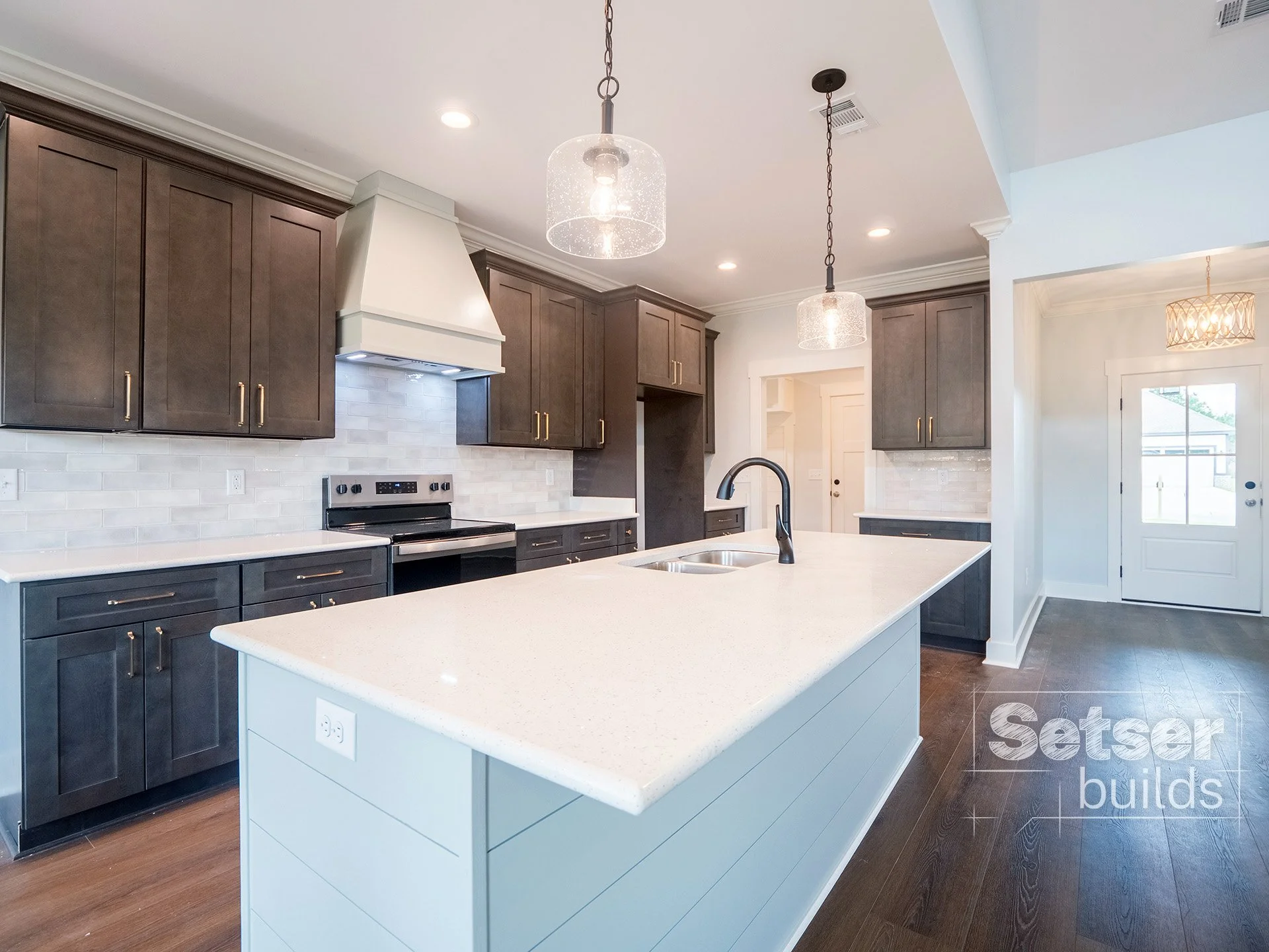 Modern kitchen with dark cabinets, white countertops, a large island with a built-in sink, pendant lights, and a stainless steel stove beneath a white range hood. There is a white brick backsplash and wood flooring.